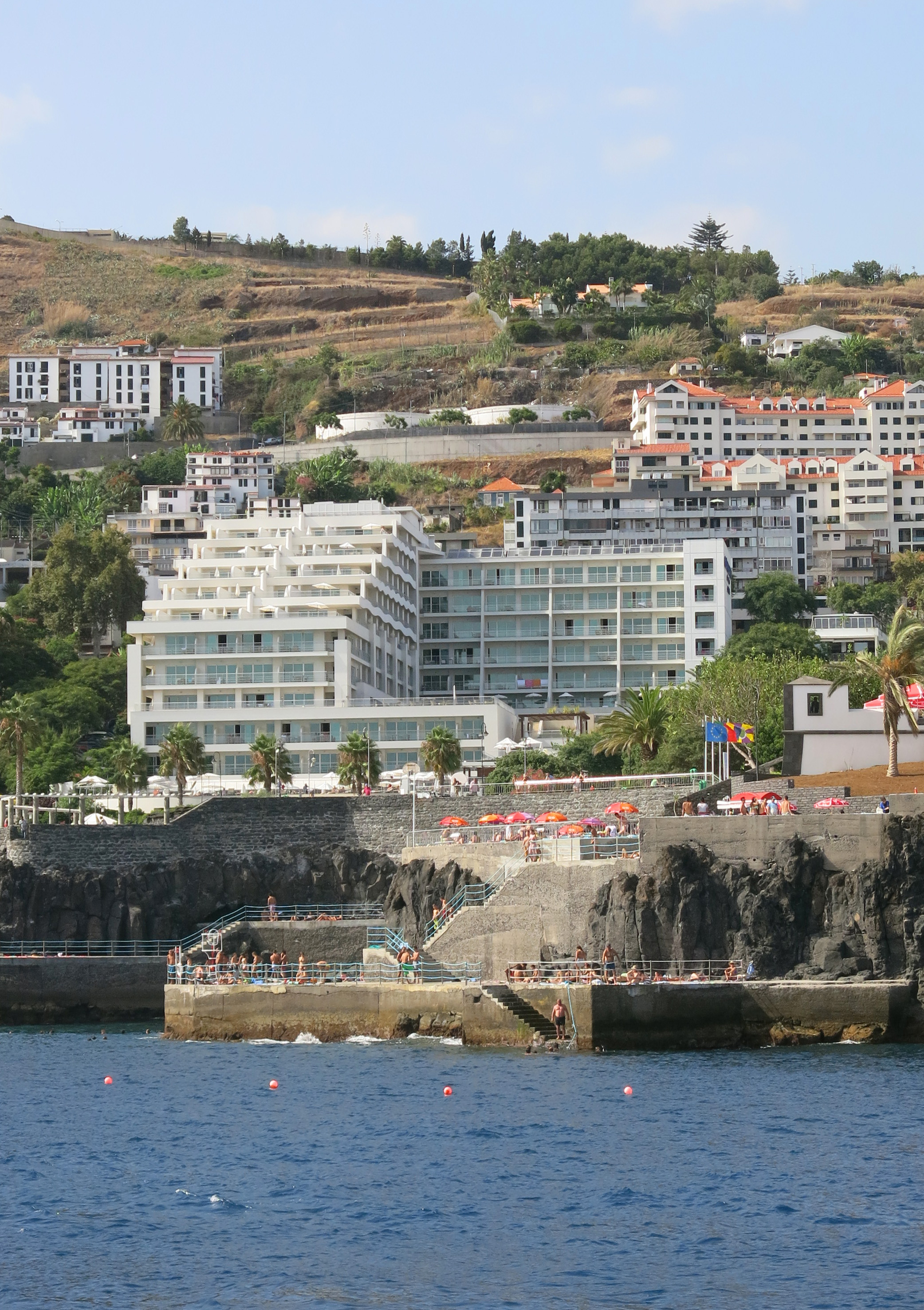 White hotels on a steep hillside overlooking rocky ocean swimming platforms with people.