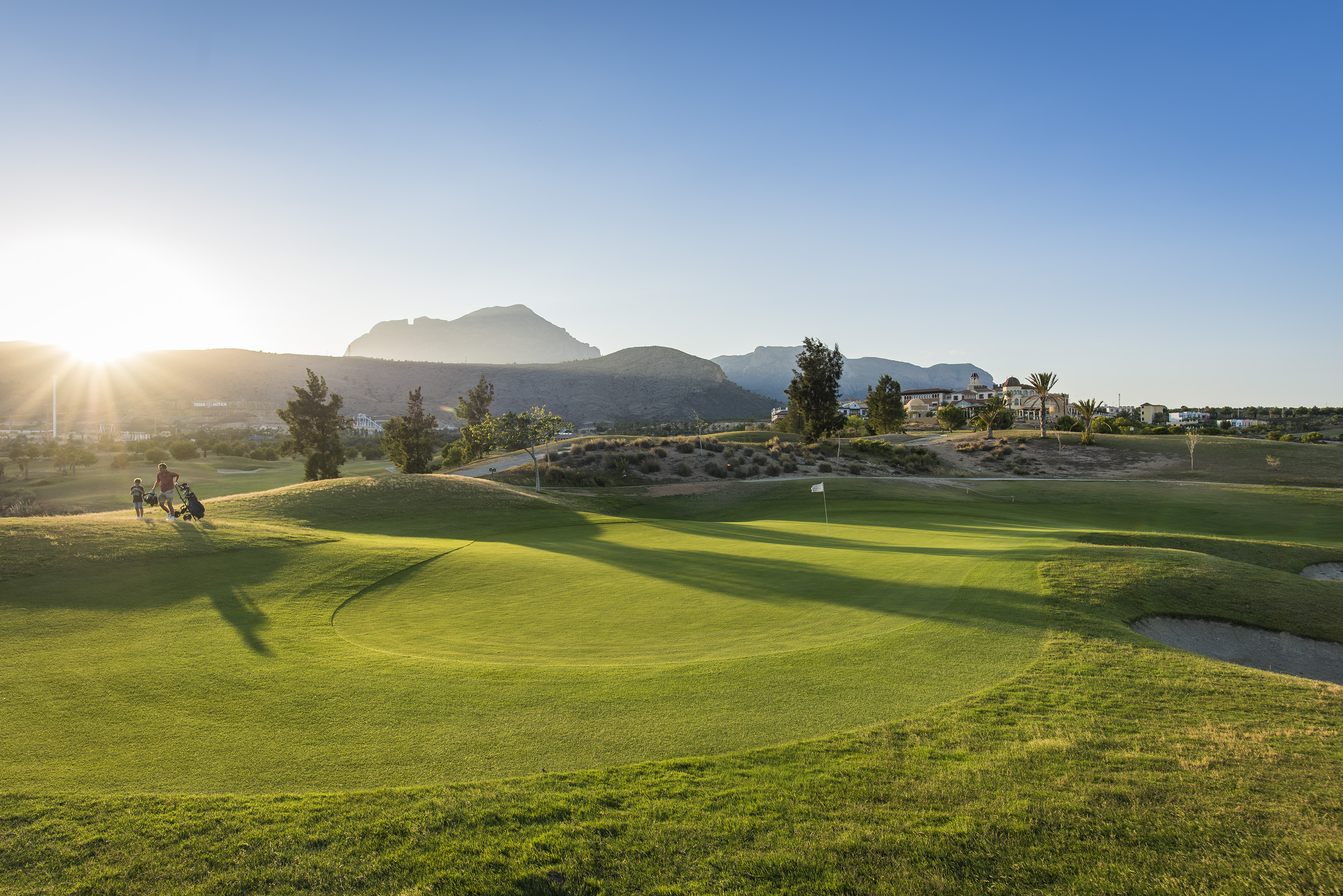 Sunset over green golf course with two golfers, mountains, and distant resort.