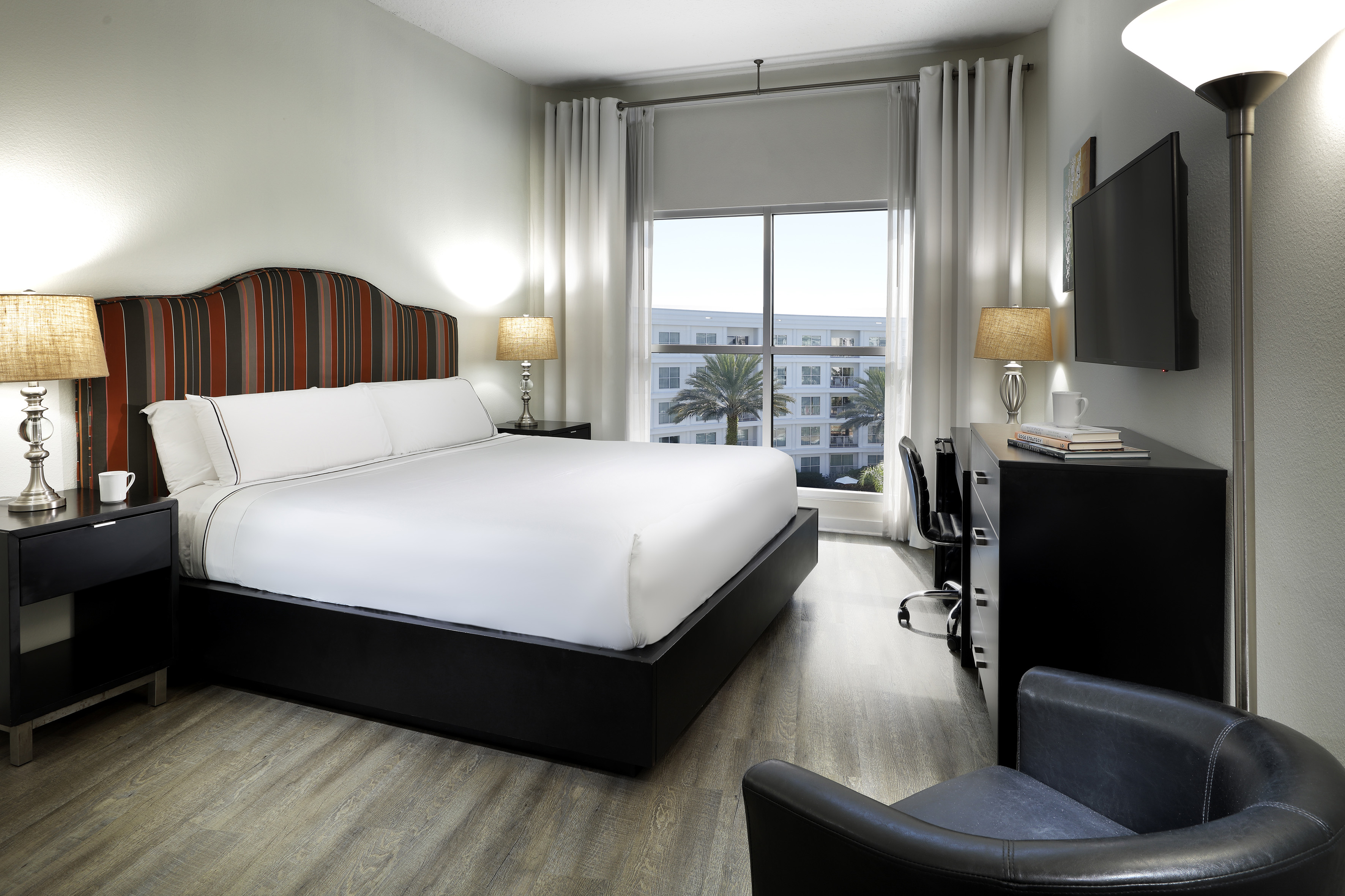 Modern hotel room with king bed, striped headboard, desk, and tropical city view. - Meliá Room - Meliá Orlando Celebration, Orlando