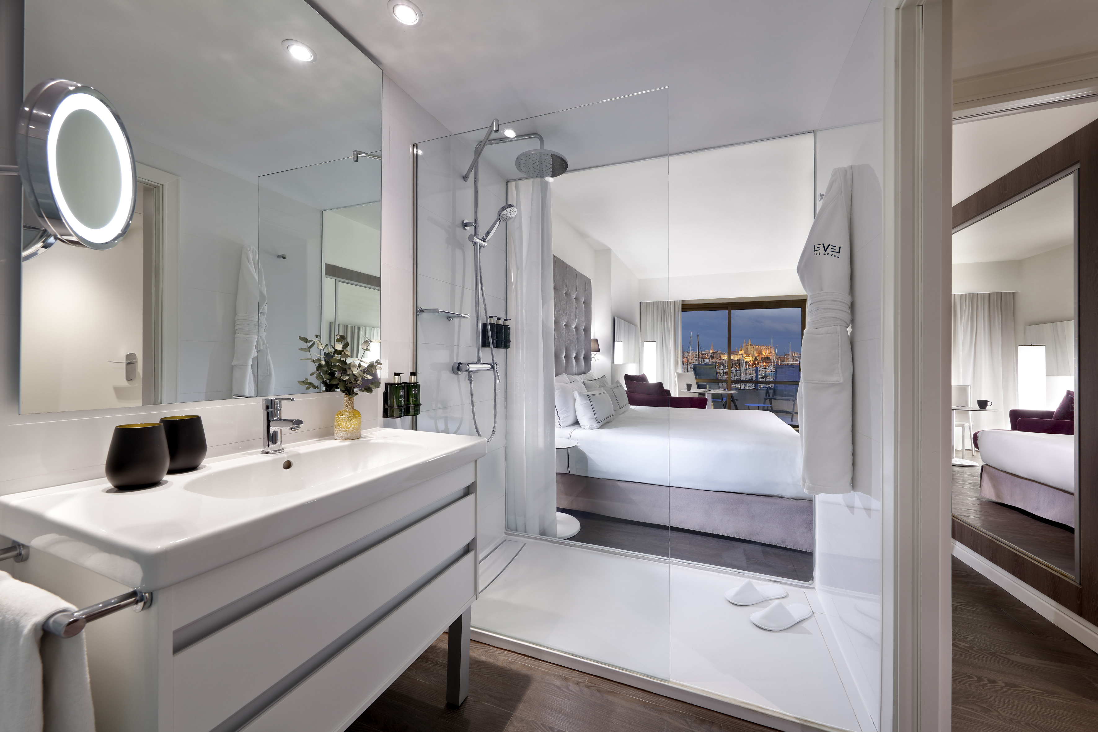 Modern hotel suite with bathroom, glass shower, and city view at dusk.