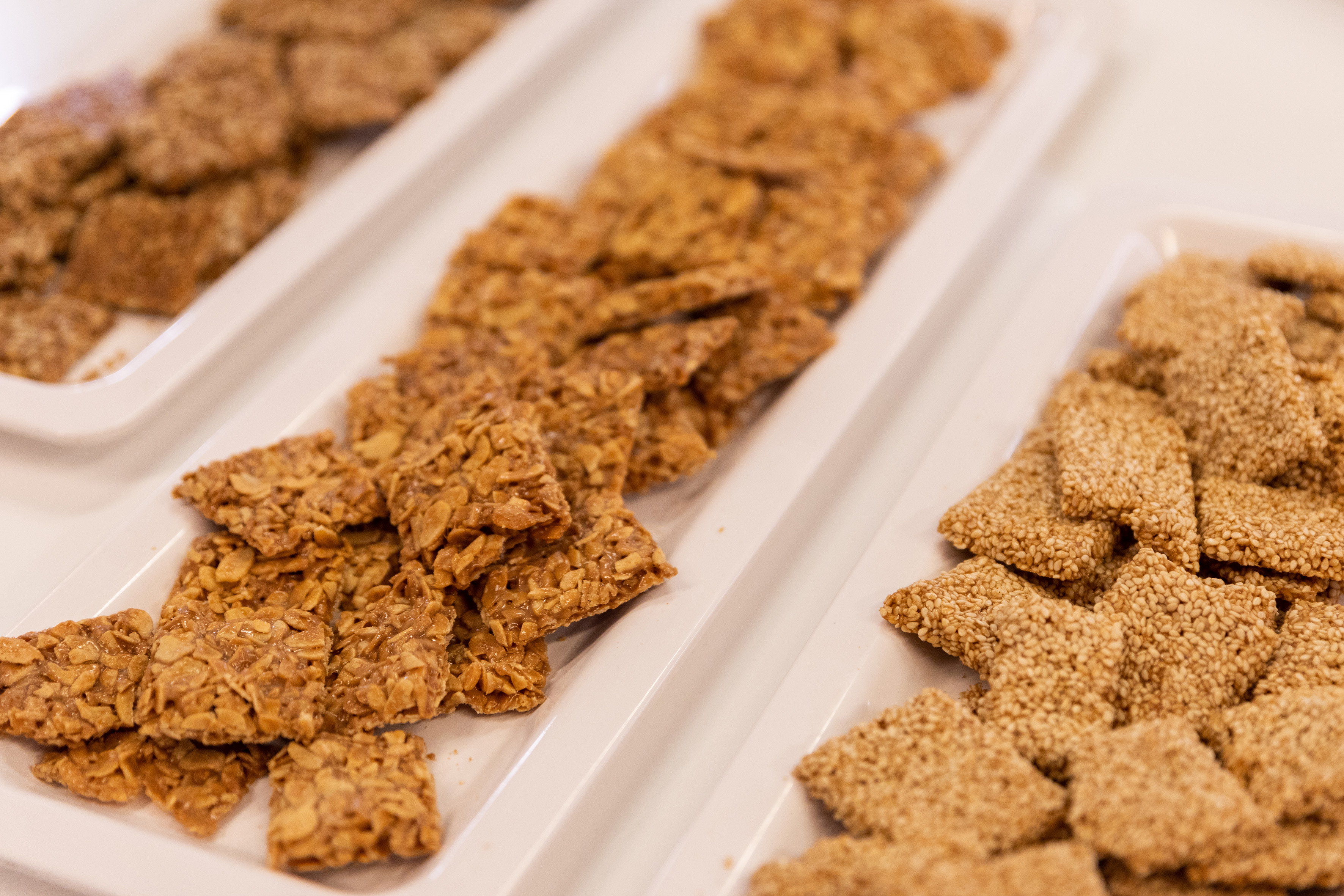 Golden almond squares and sesame brittle on white serving trays.
