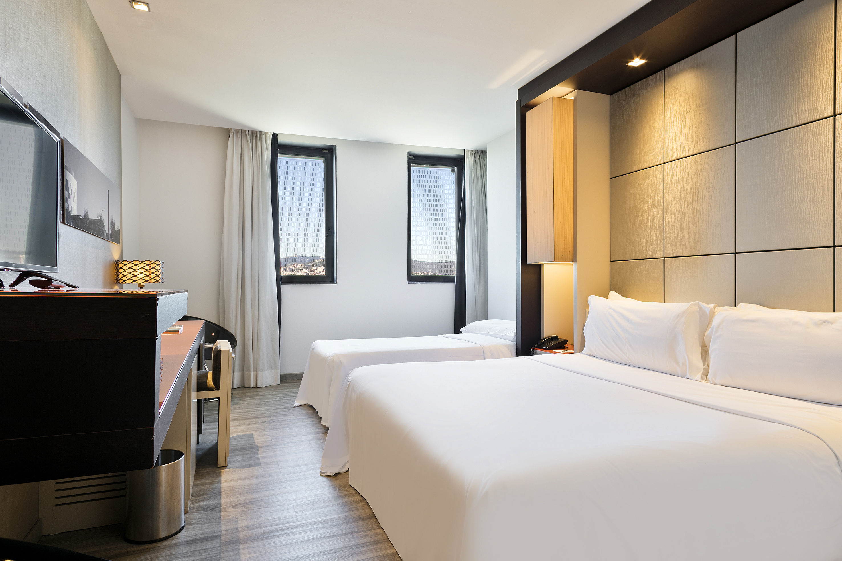 Modern hotel room with queen bed, twin bed, city view windows, and desk.