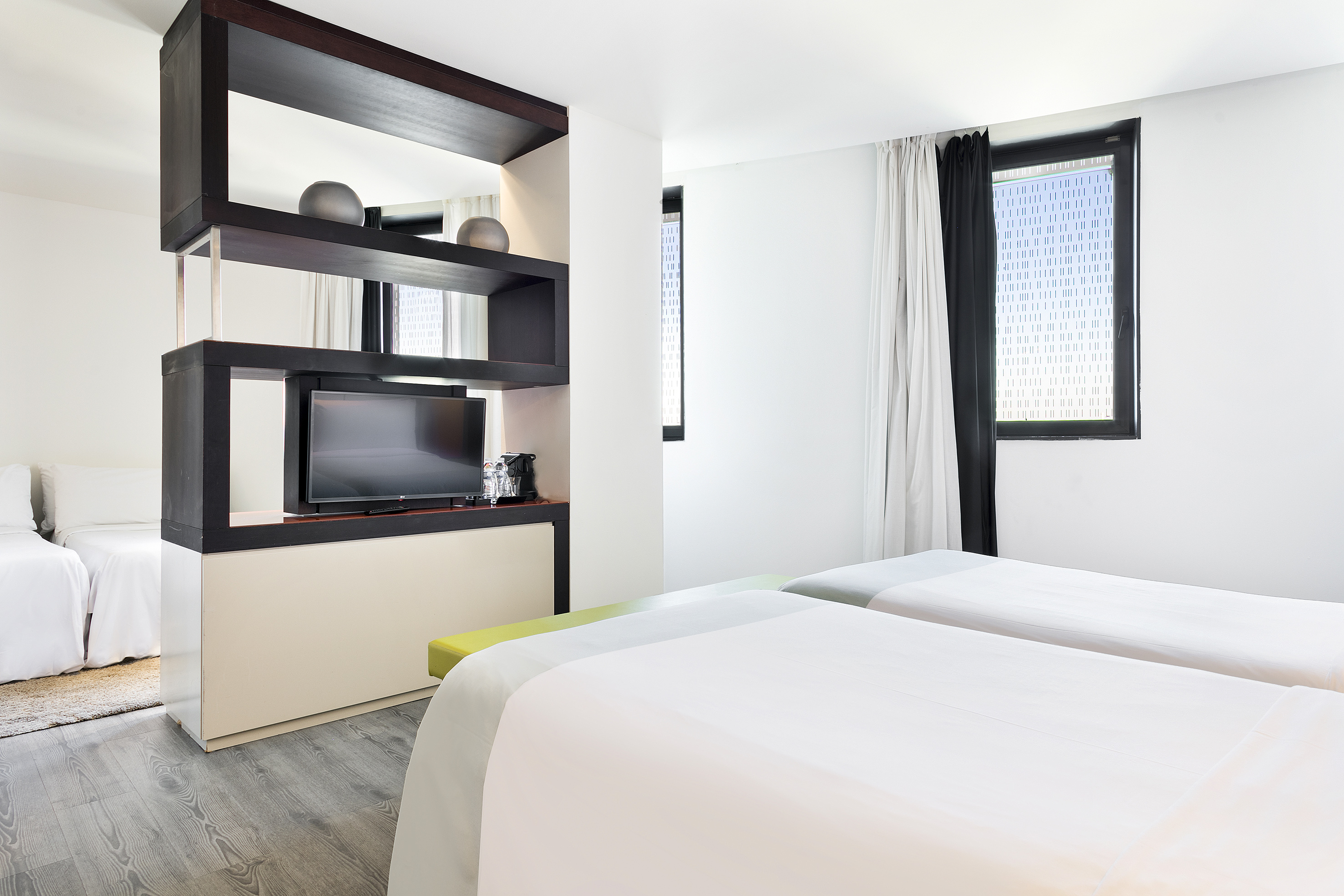 Modern hotel bedroom with twin beds, wood room divider, TV, and window.