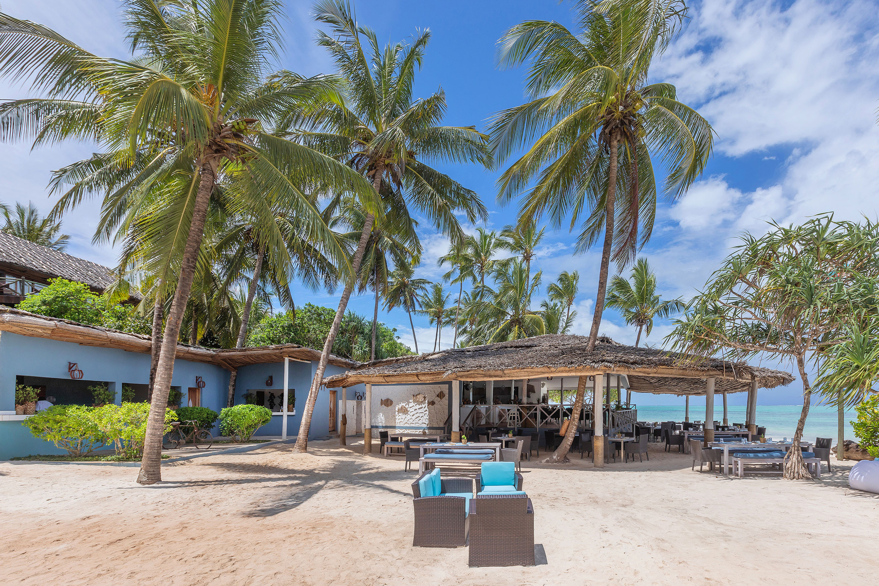 Beachfront dining area with thatched roof, palm trees, and sandy beach.