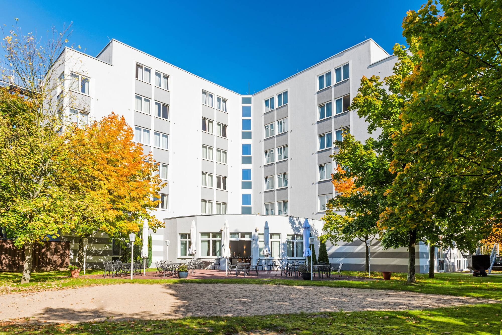 Modern hotel building with outdoor dining terrace, vibrant autumn trees, and blue sky.