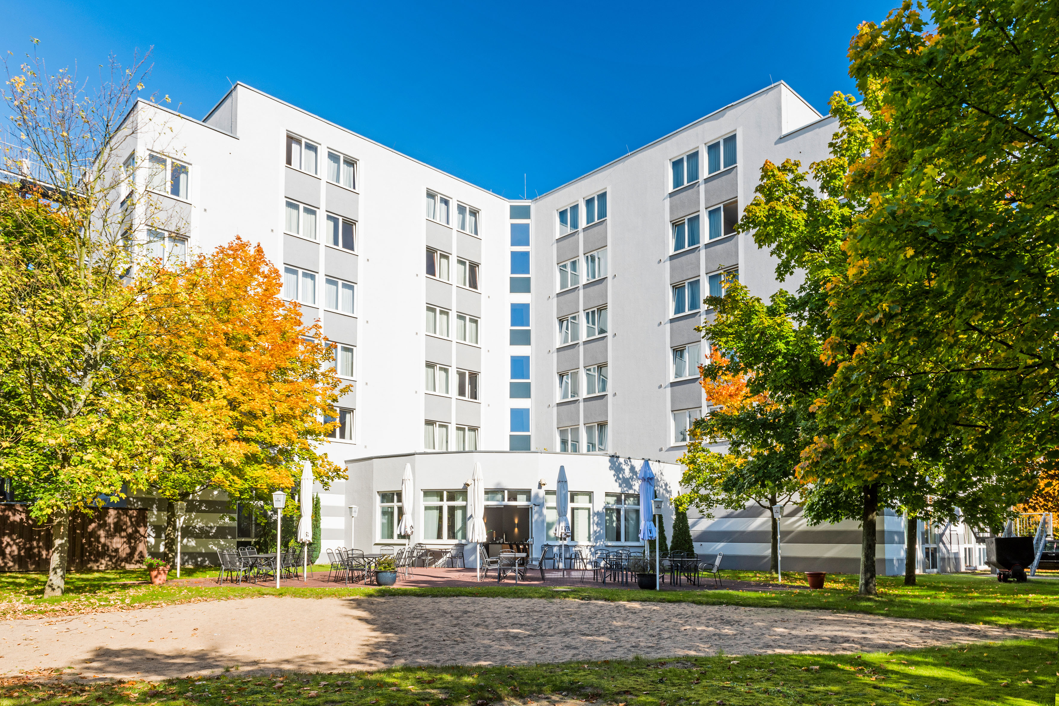 Modern hotel building with outdoor dining terrace, vibrant autumn trees, and blue sky.