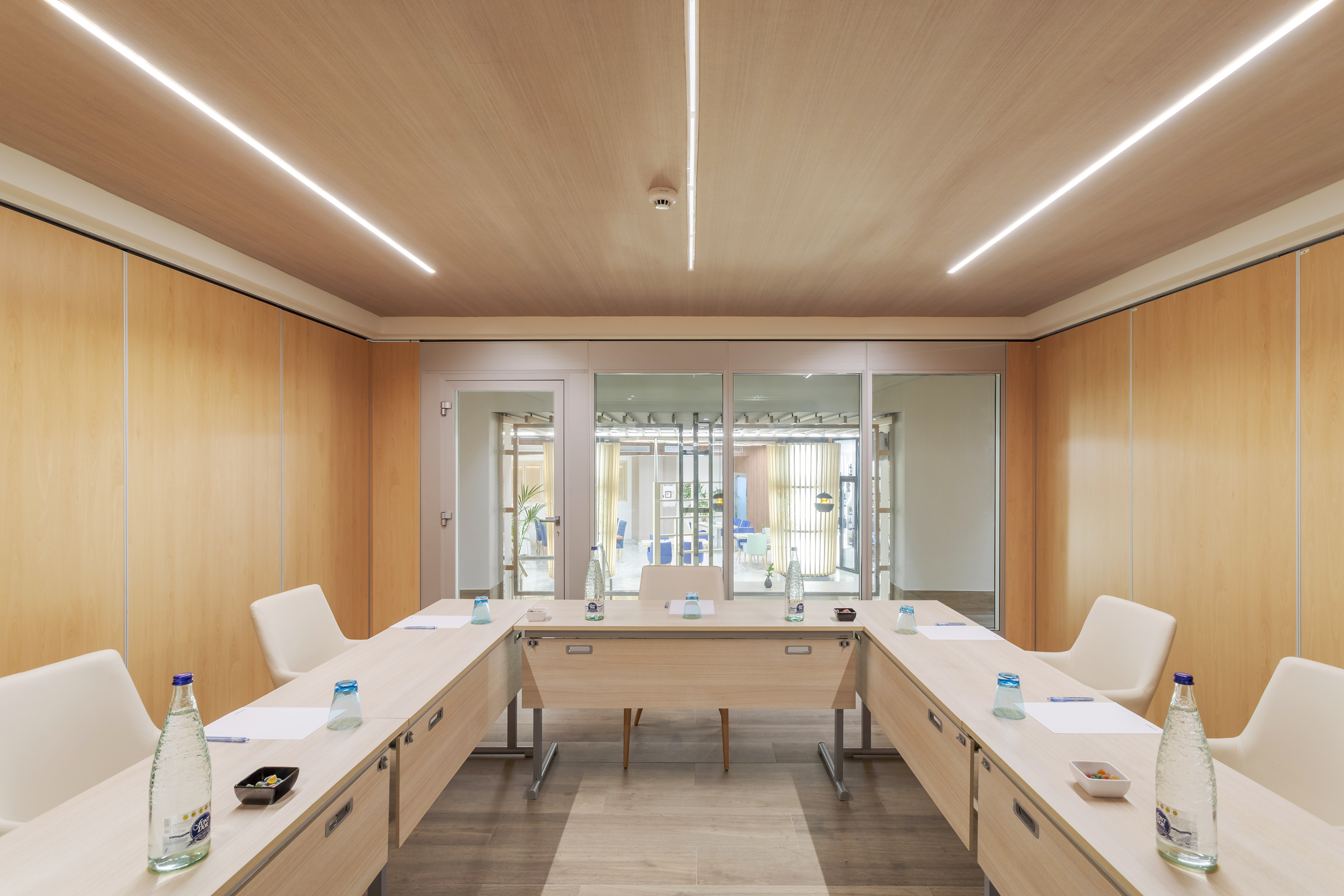 Modern conference room with U-shaped wood table, linear LED lights, and glass walls.