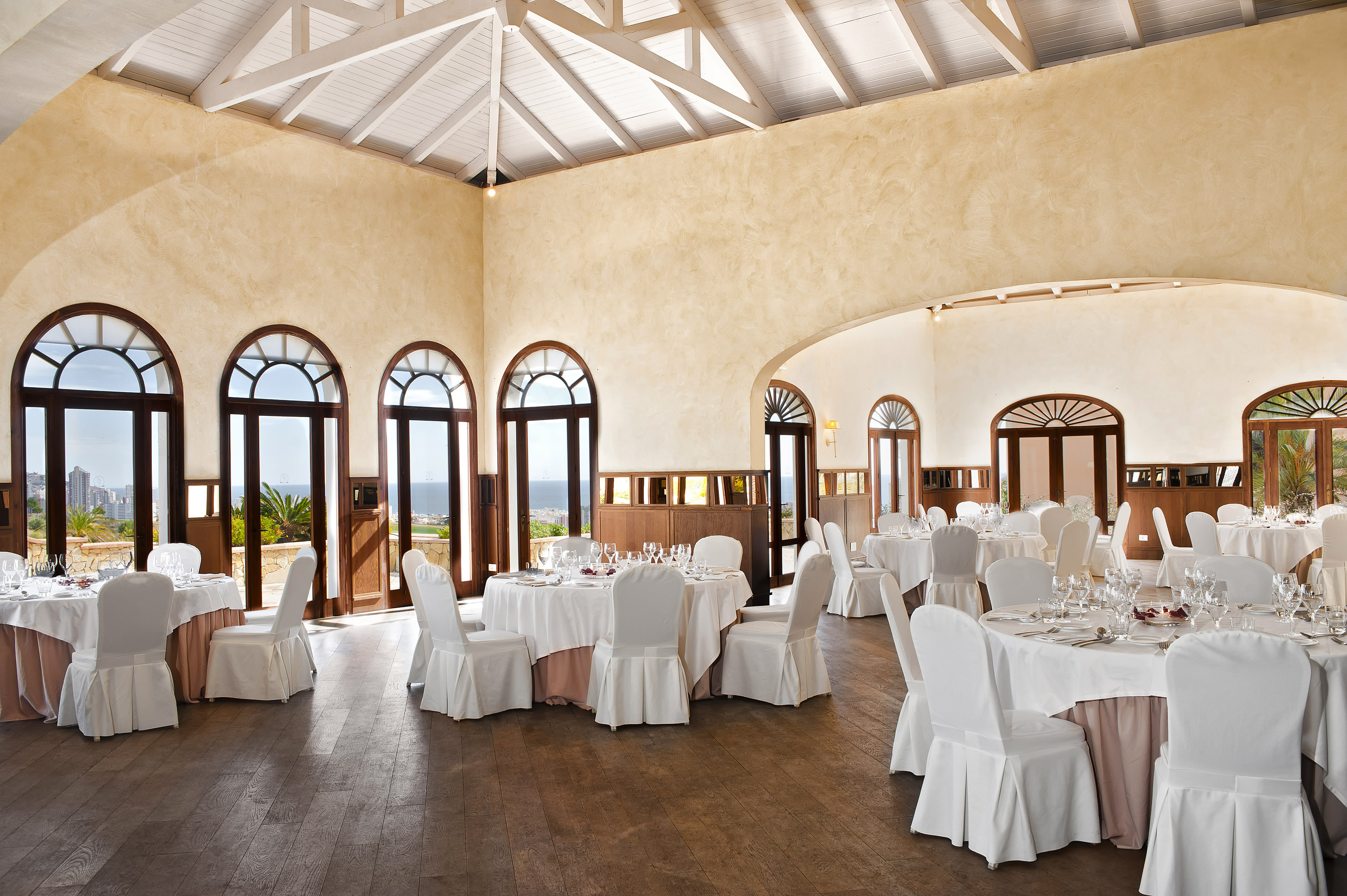 Formal dining hall with white tables, arched windows, and coastal view.