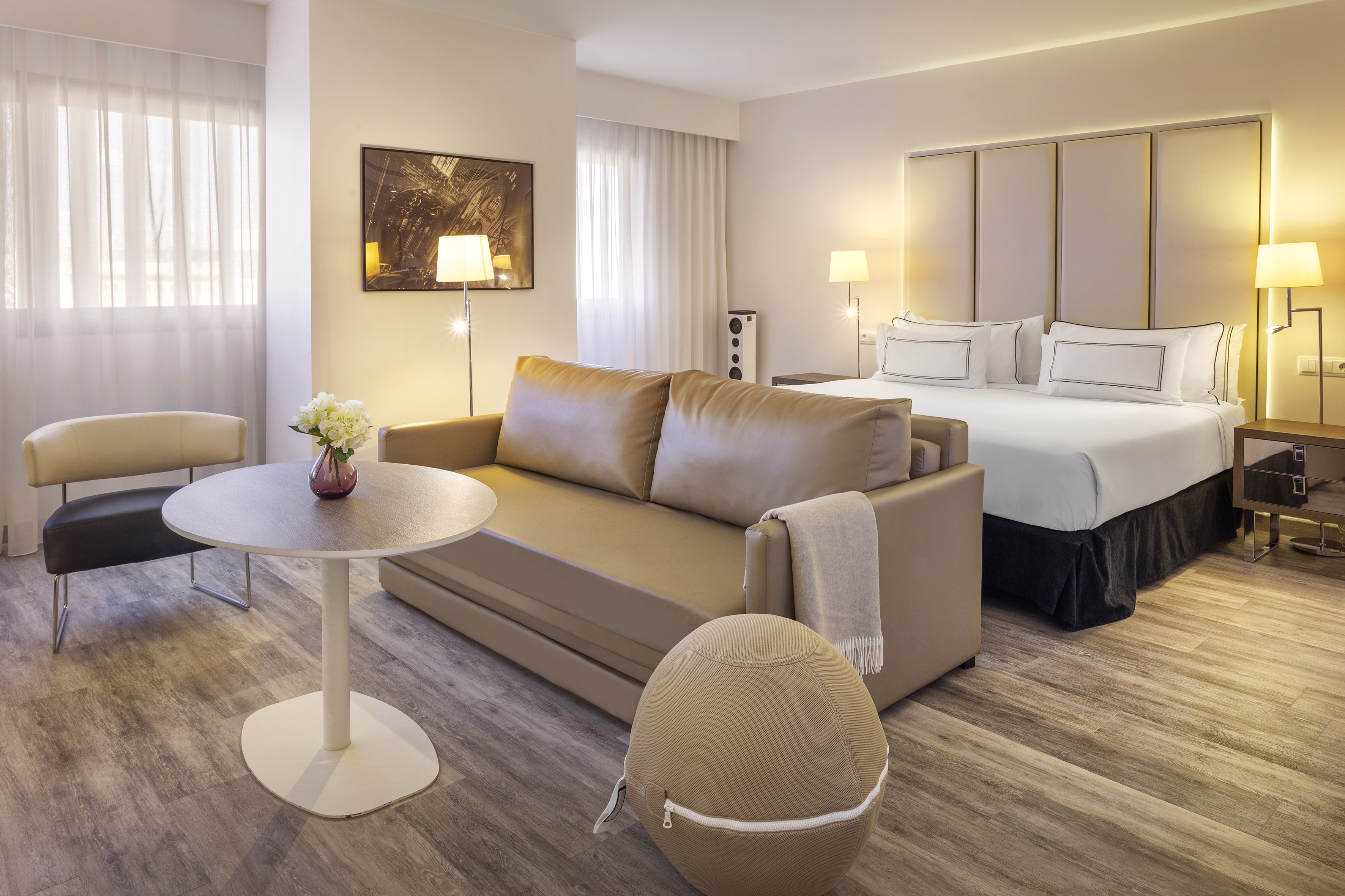 Modern hotel room with king bed, tan sofa, wooden floor, and sheer curtains.
