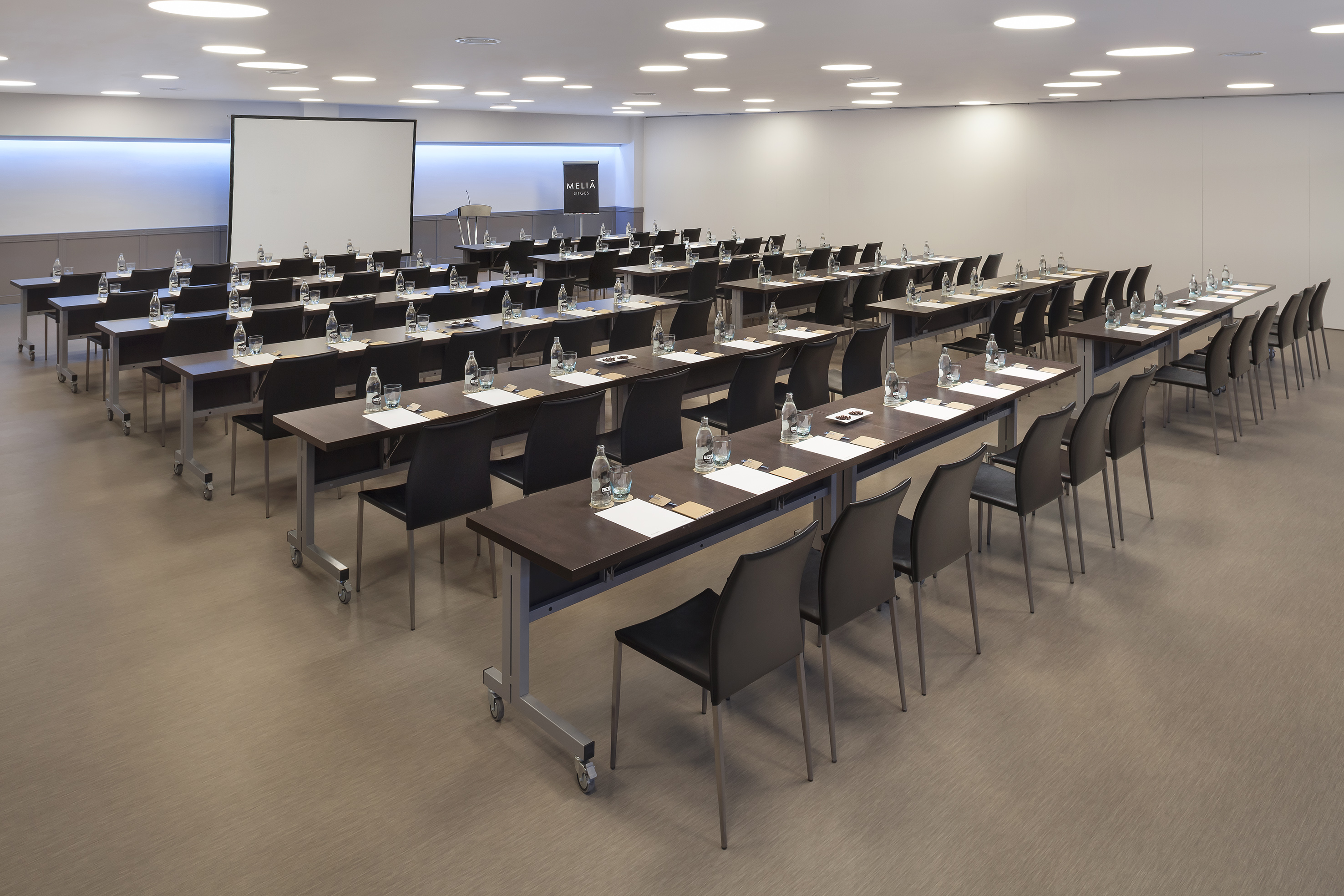 Hotel conference room setup with tables, chairs, water bottles, and projection screen.