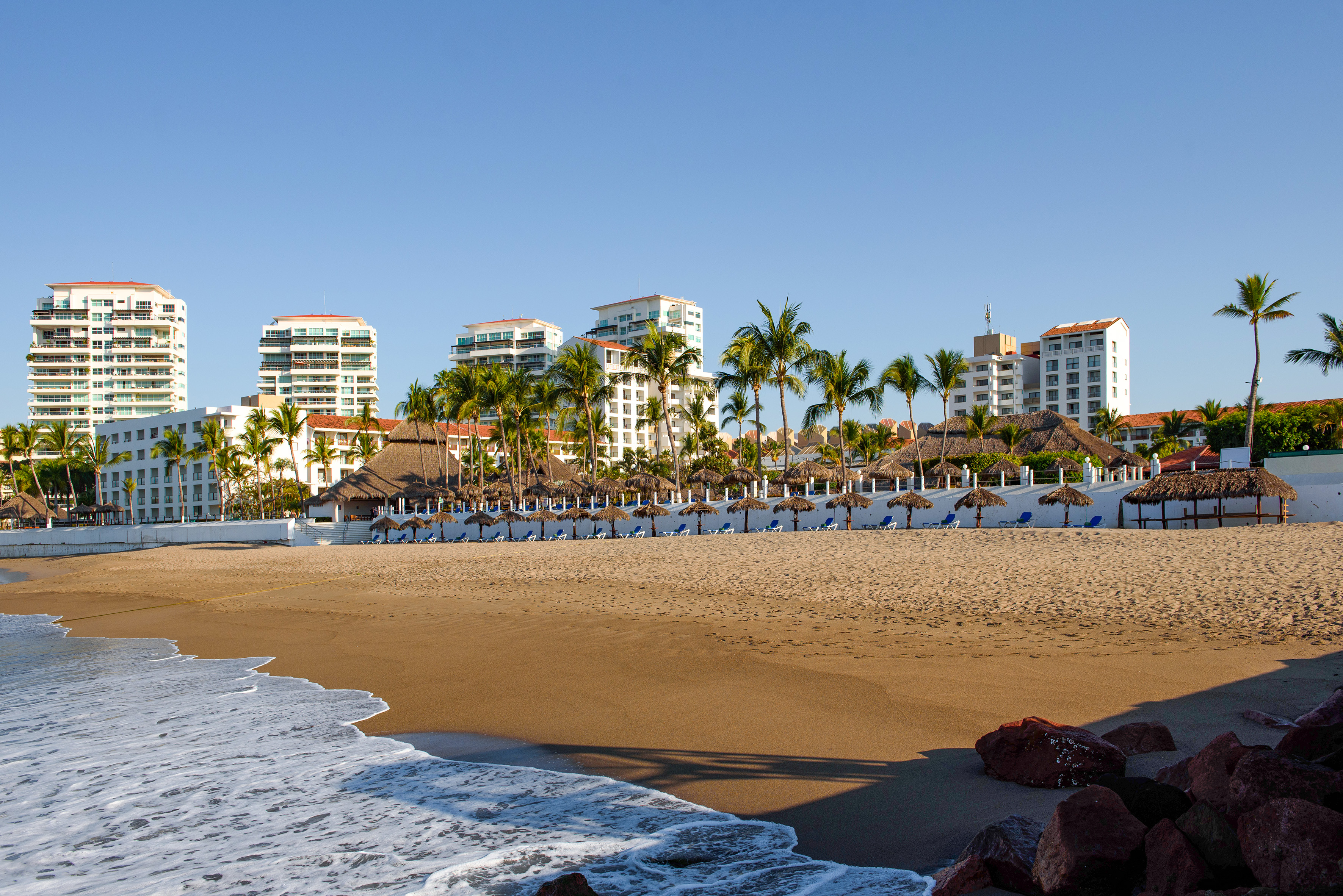 Tropical resort beach with palm trees, thatched umbrellas, and ocean waves.