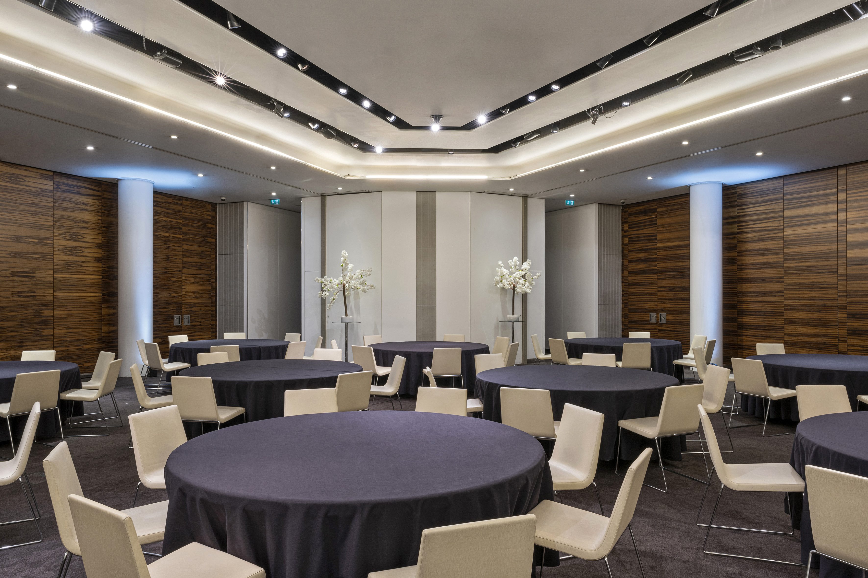 Modern meeting room with round tables, dark linens, light chairs, and wood paneling.