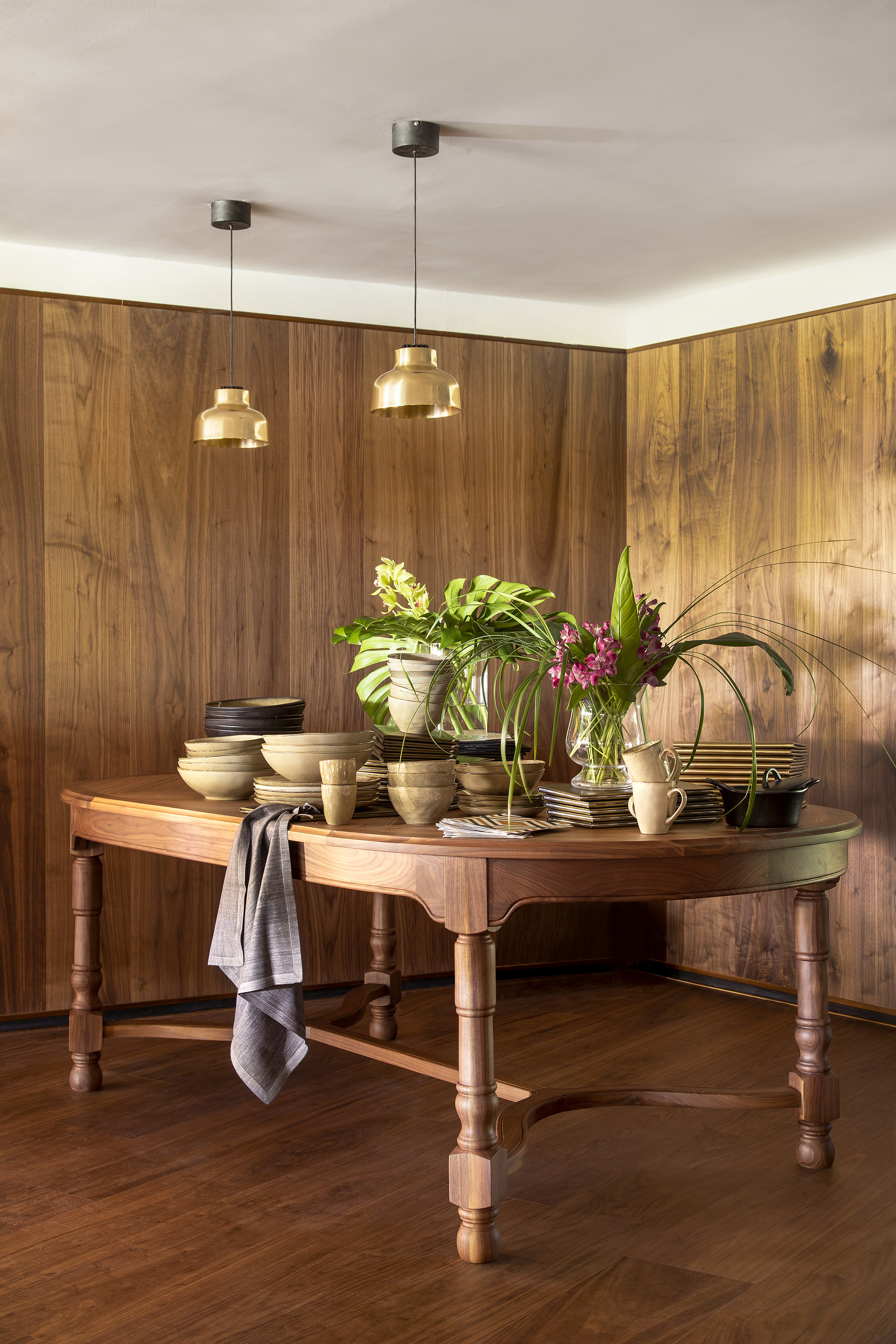 Wooden dining table with rustic dinnerware, flowers, brass lights, wood walls.