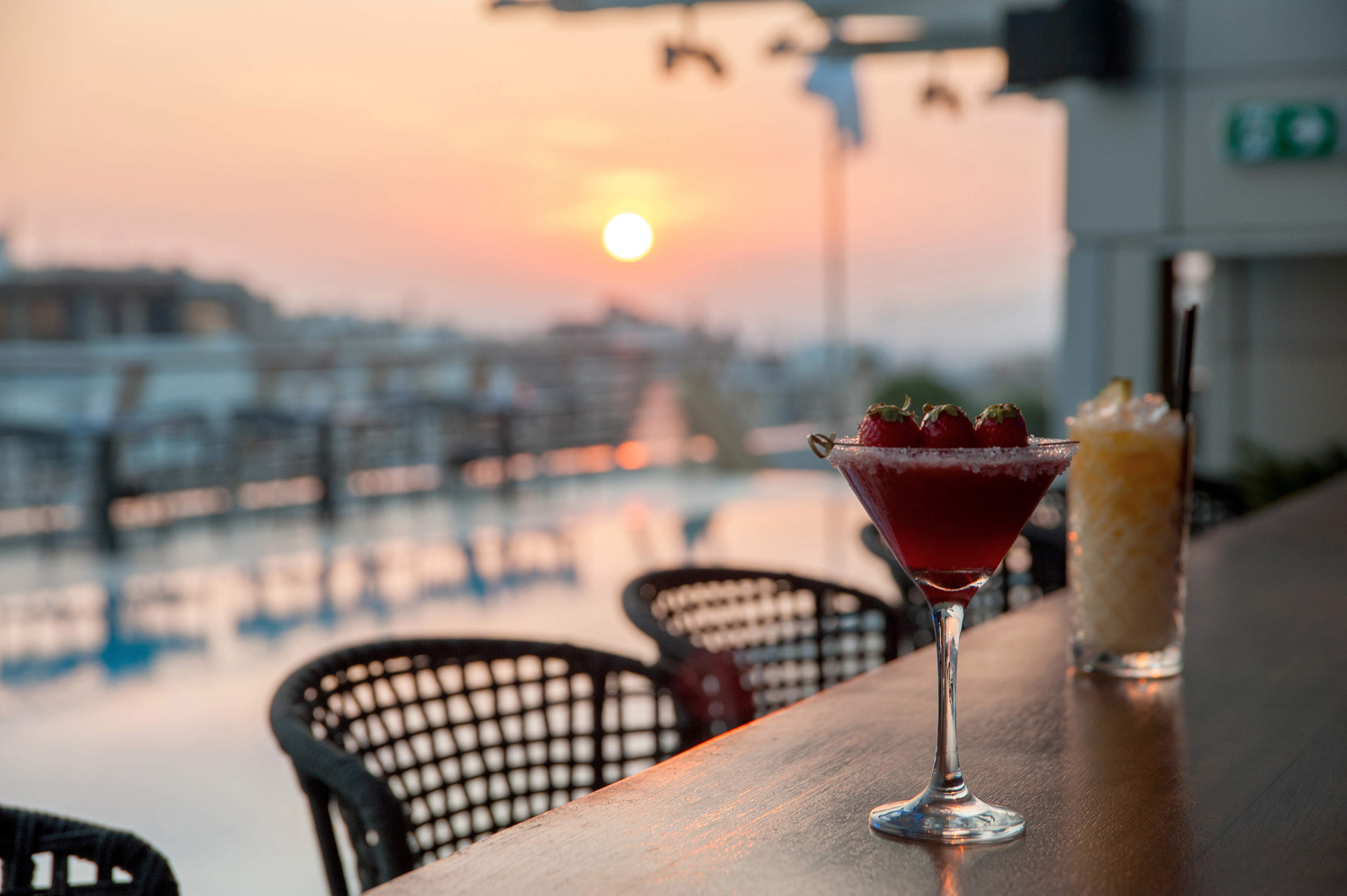 Red cocktail with strawberries on a bar at sunset over a marina.