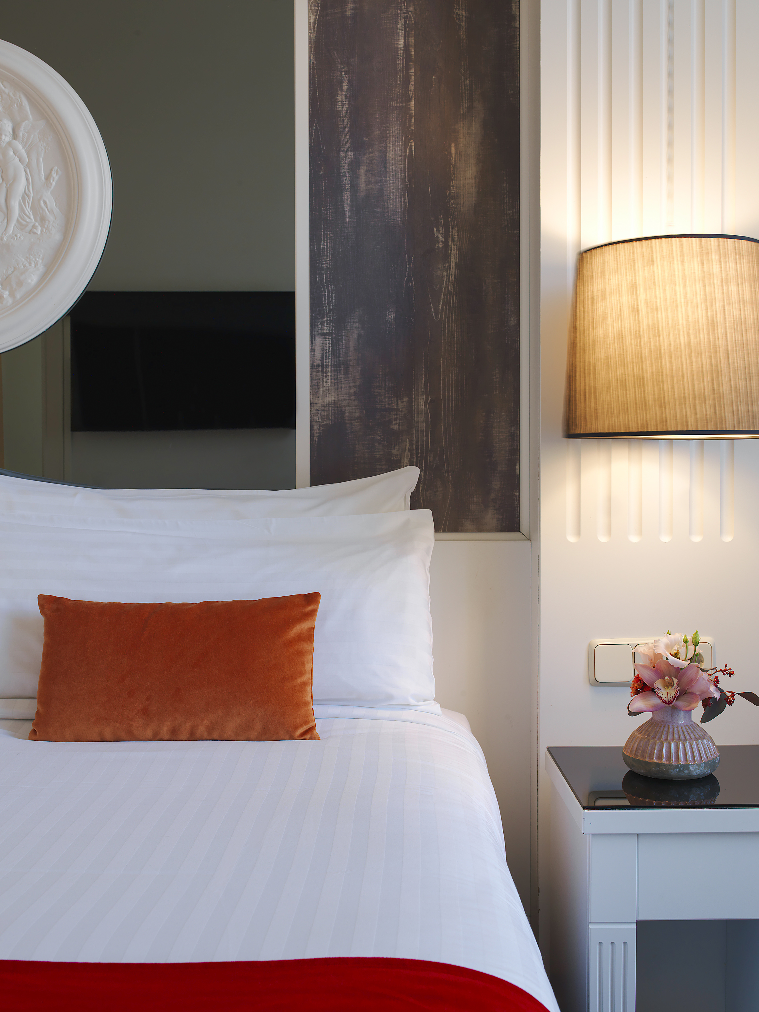 Contemporary hotel room bed with white sheets, orange pillow, and wall lamp.