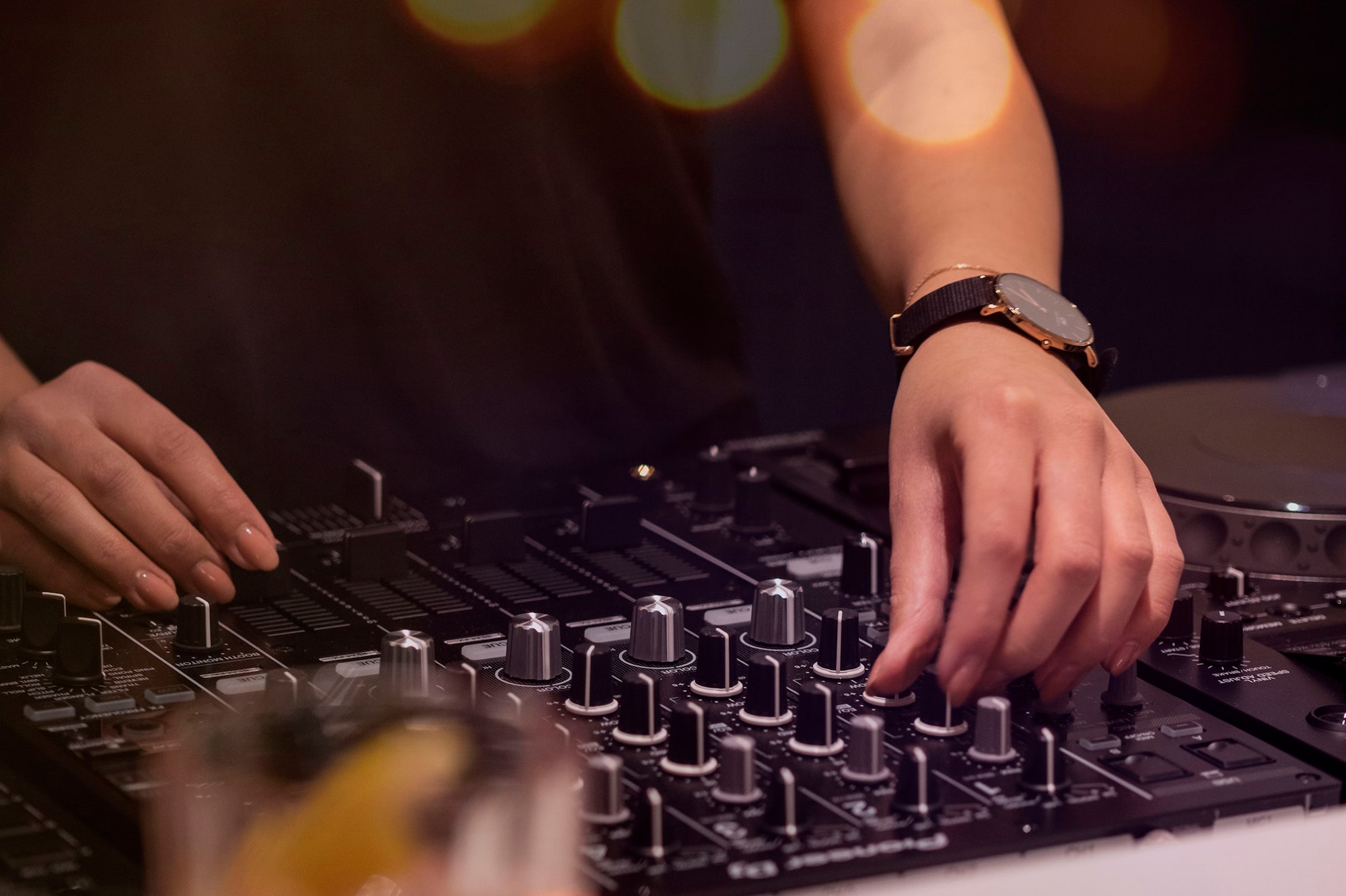 DJ mixing music with hands on a controller, cocktail in foreground, bokeh lights.