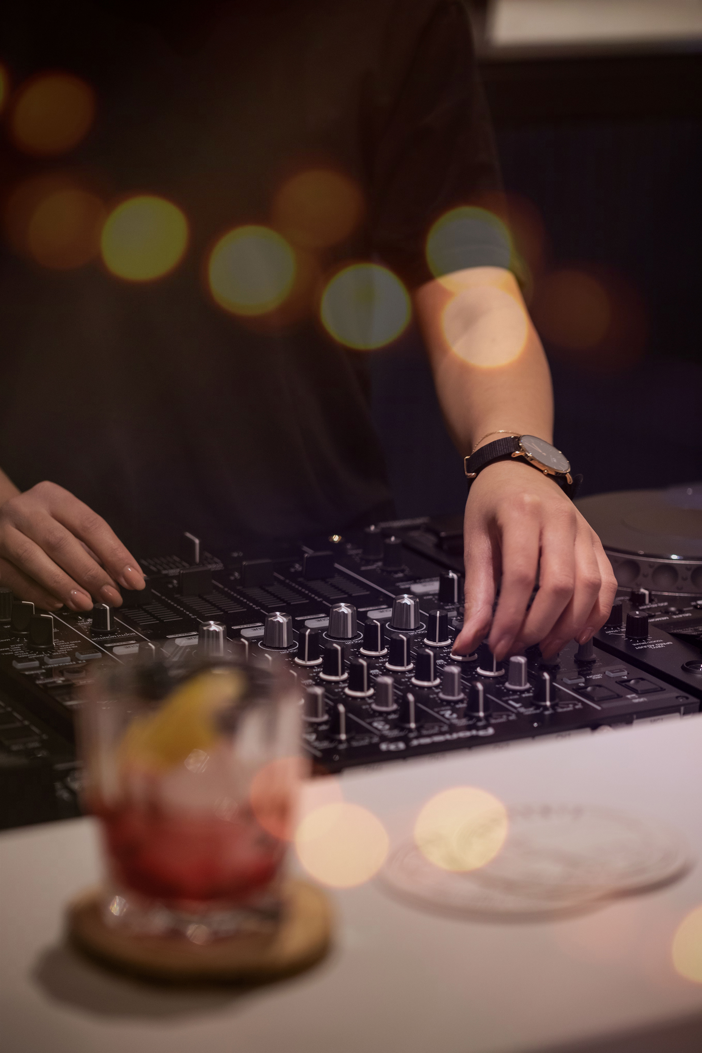 DJ mixing music with hands on a controller, cocktail in foreground, bokeh lights.