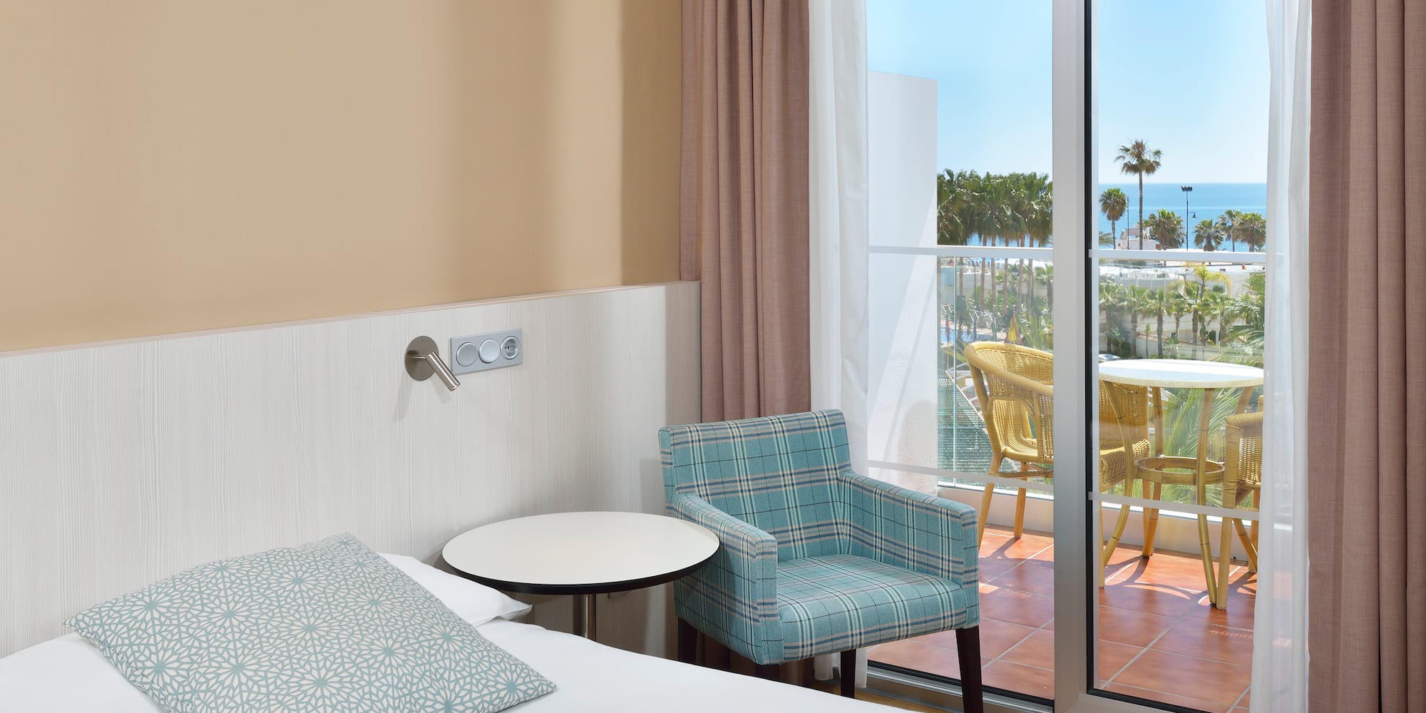 Hotel room with bed, armchair, and balcony with ocean view and palm trees.