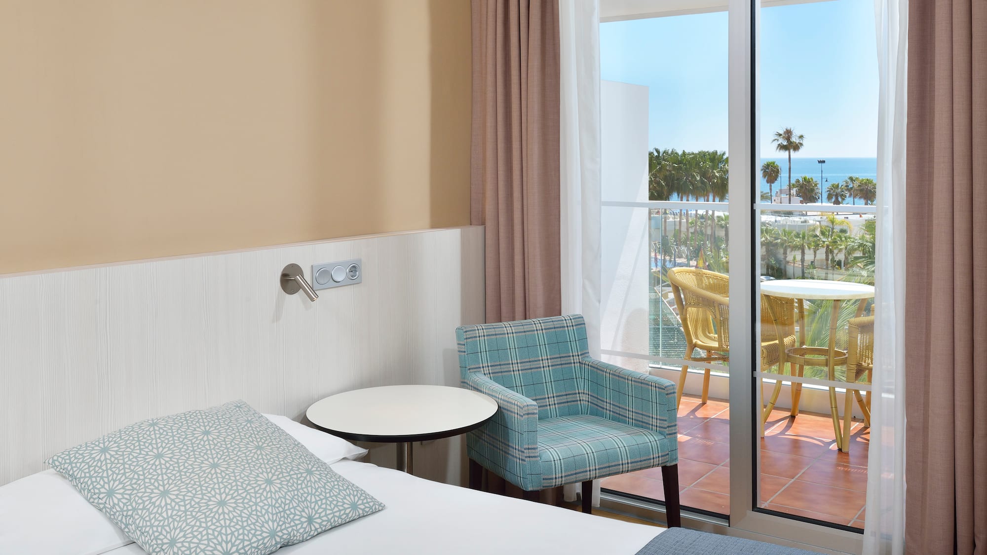 Hotel room with bed, armchair, and balcony with ocean view and palm trees.