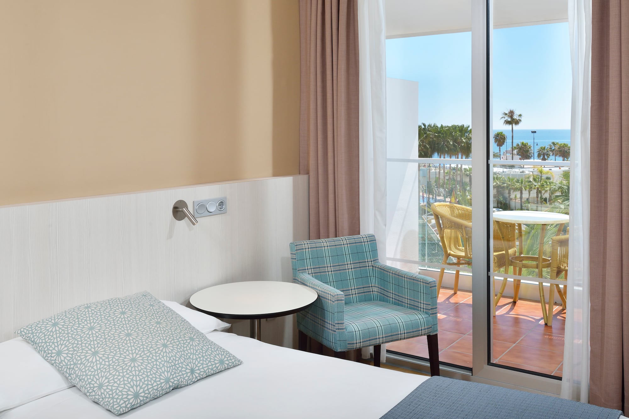 Hotel room with bed, armchair, and balcony with ocean view and palm trees.