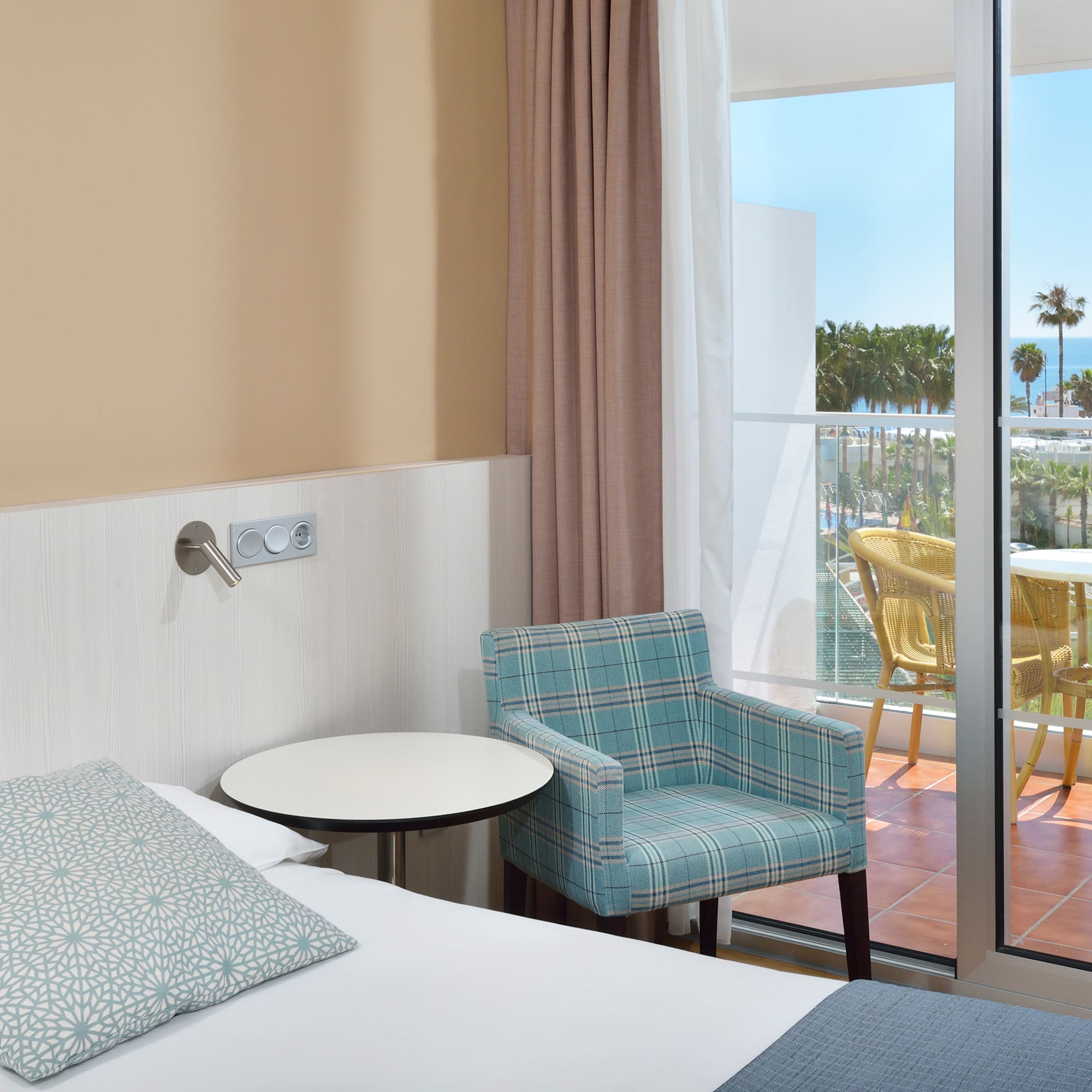 Hotel room with bed, armchair, and balcony with ocean view and palm trees.