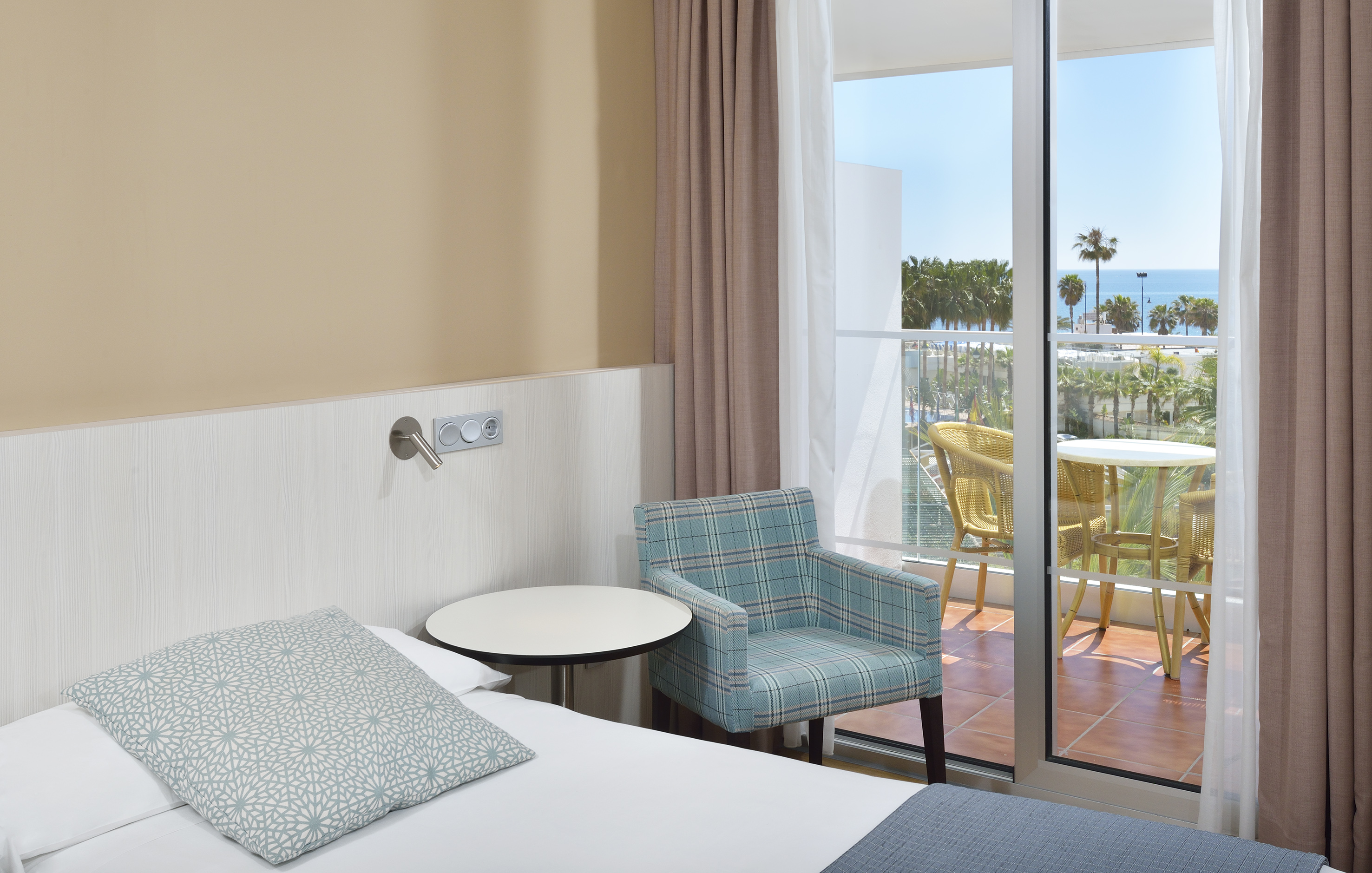 Hotel room with bed, armchair, and balcony with ocean view and palm trees.