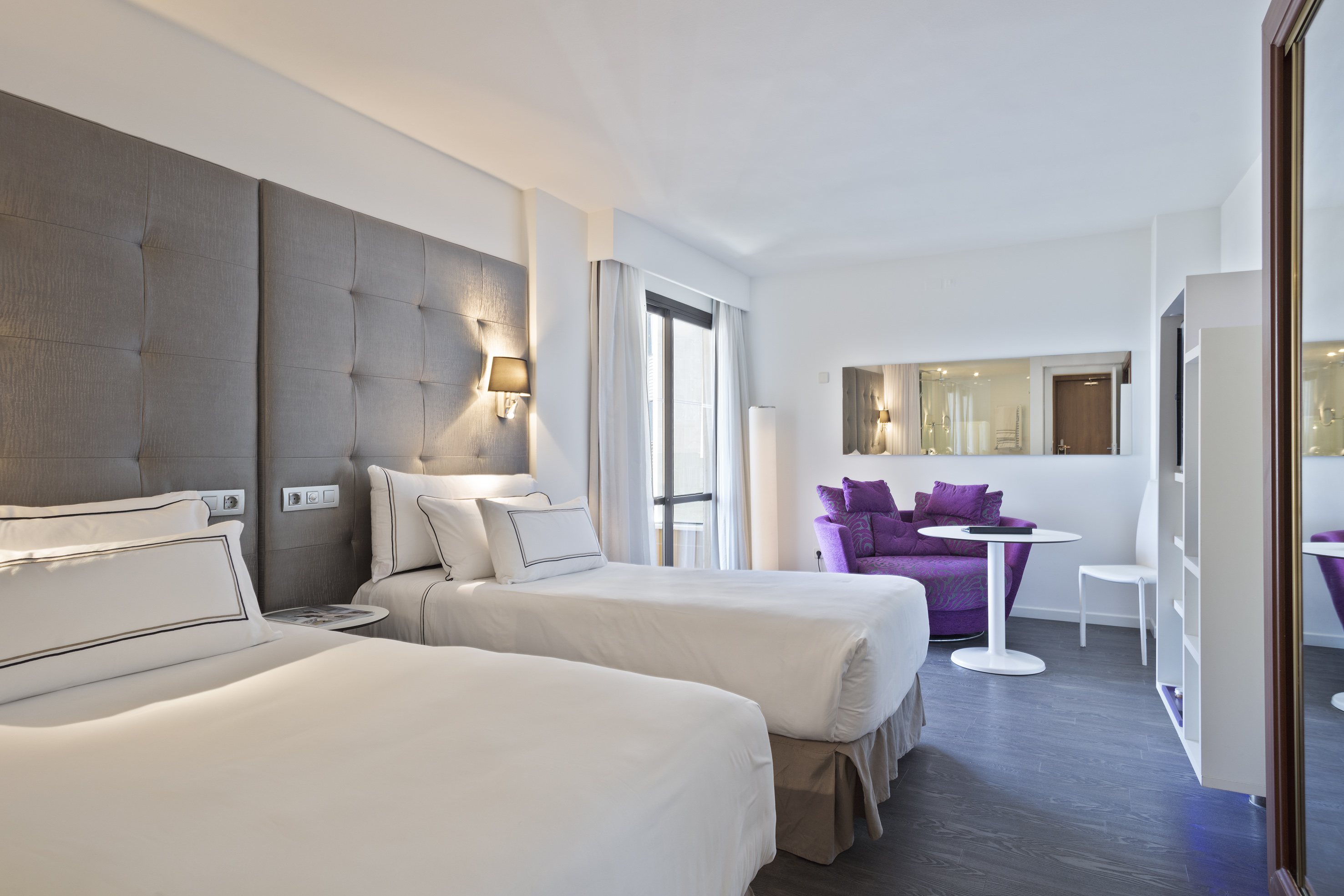 Modern hotel guest room with twin beds, grey headboards, and a vibrant purple sofa.