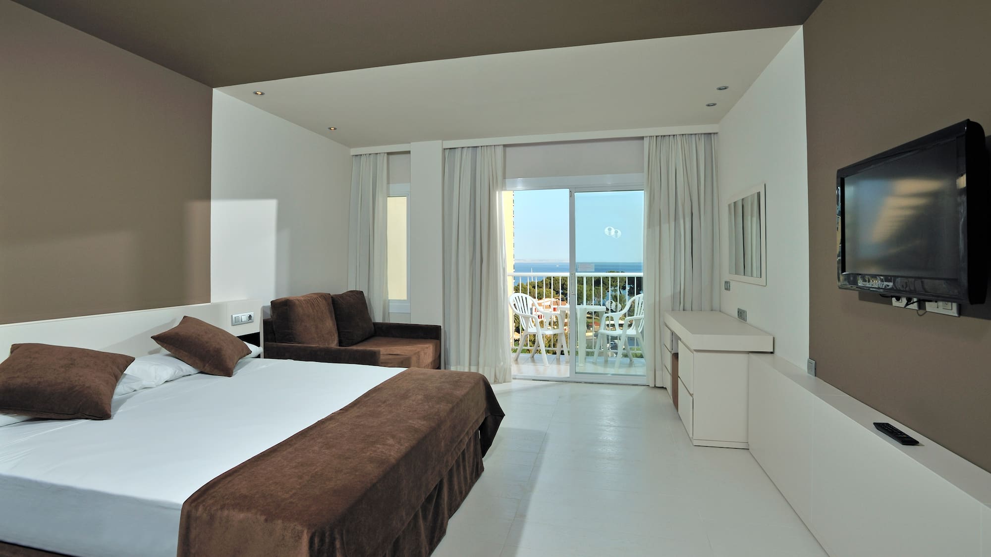 Modern hotel room with sea view balcony, white bed, and brown sofa.