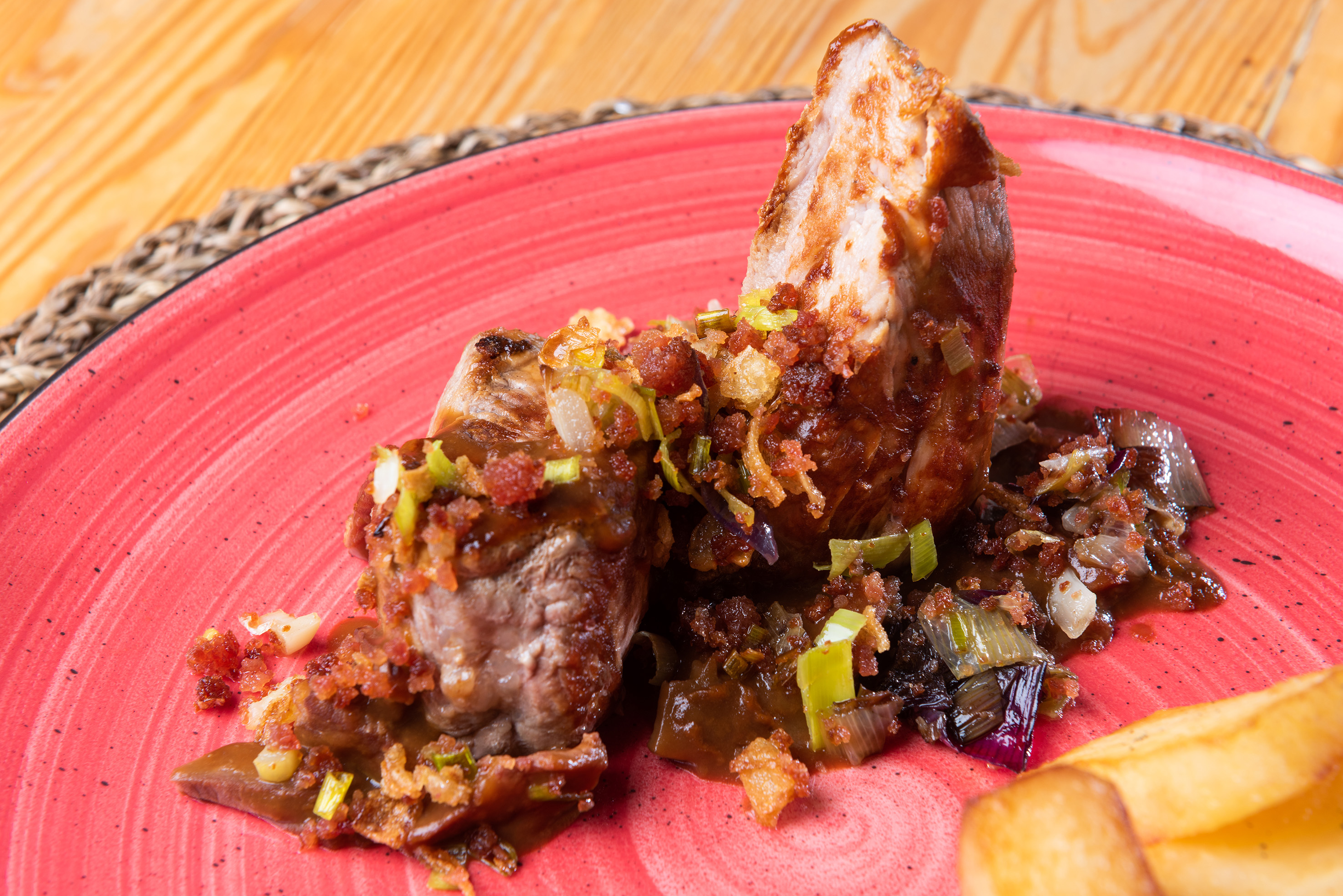 Pork loin with gravy, crispy bacon, green onions, and fries on a red plate.