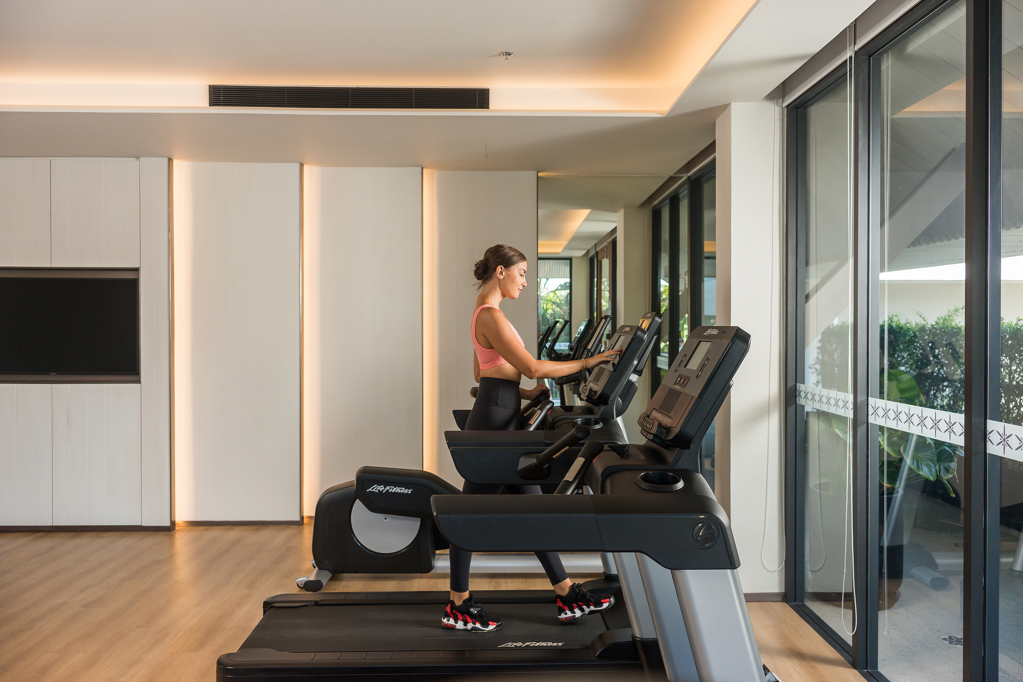 Woman exercising on a treadmill in a bright, modern gym with large windows.