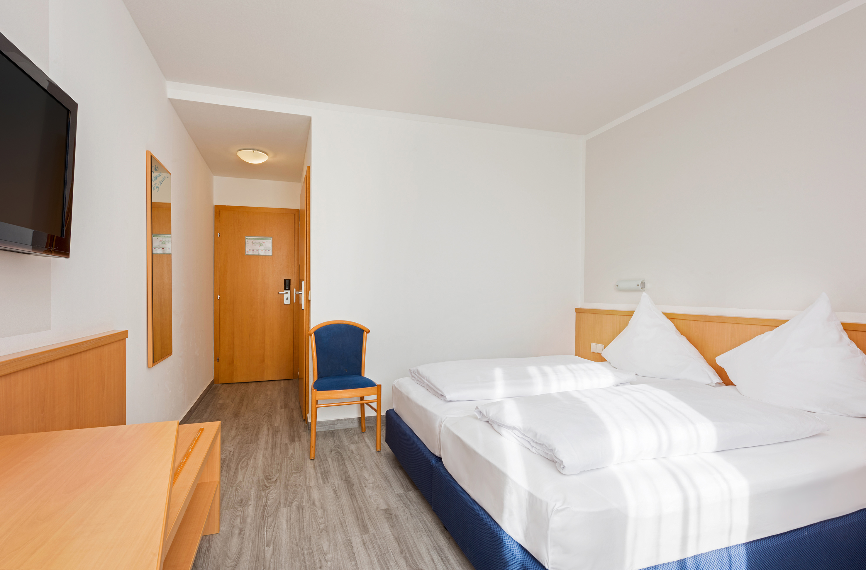 Bright hotel room with twin beds, blue chair, wooden door, and wall-mounted TV.