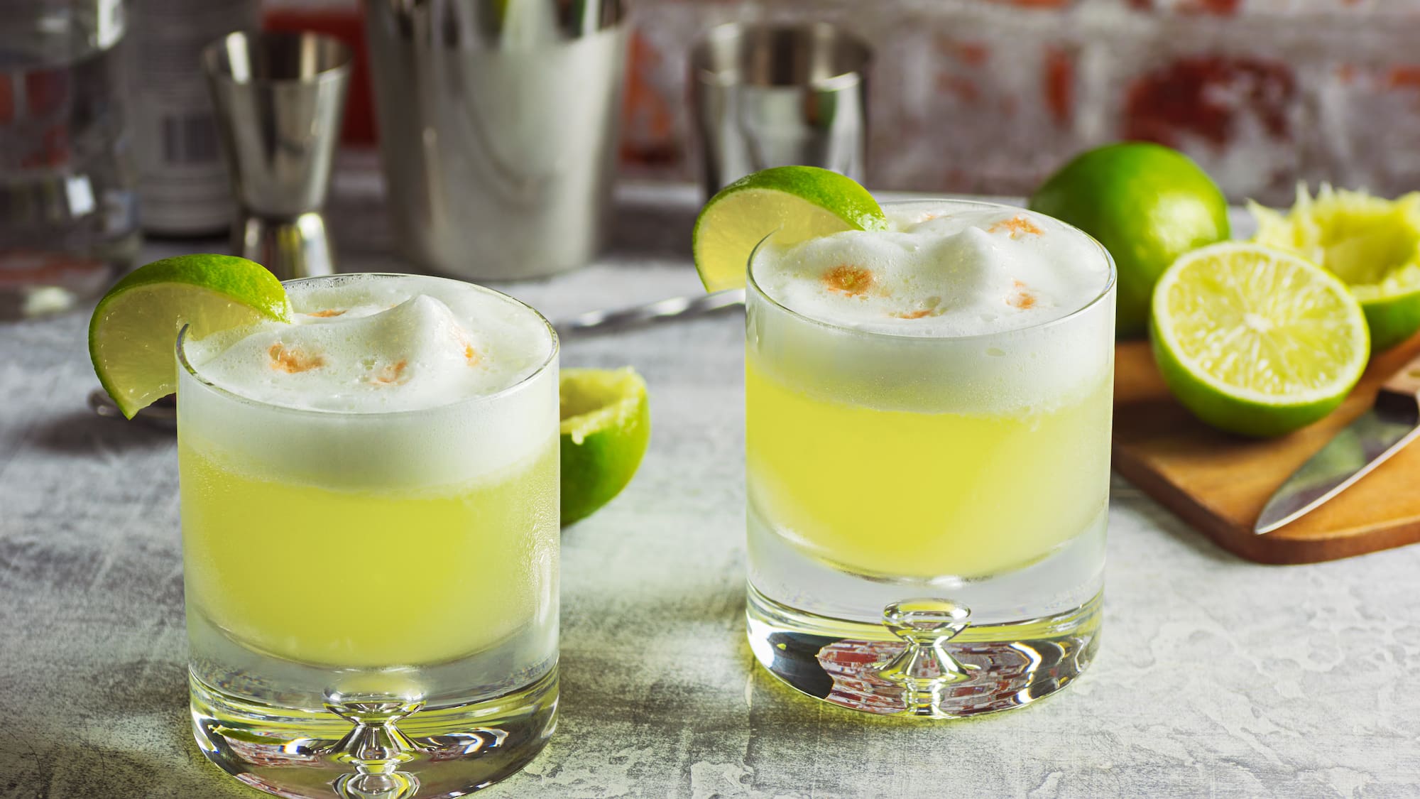 two glasses of yellow liquid with limes and a cutting board