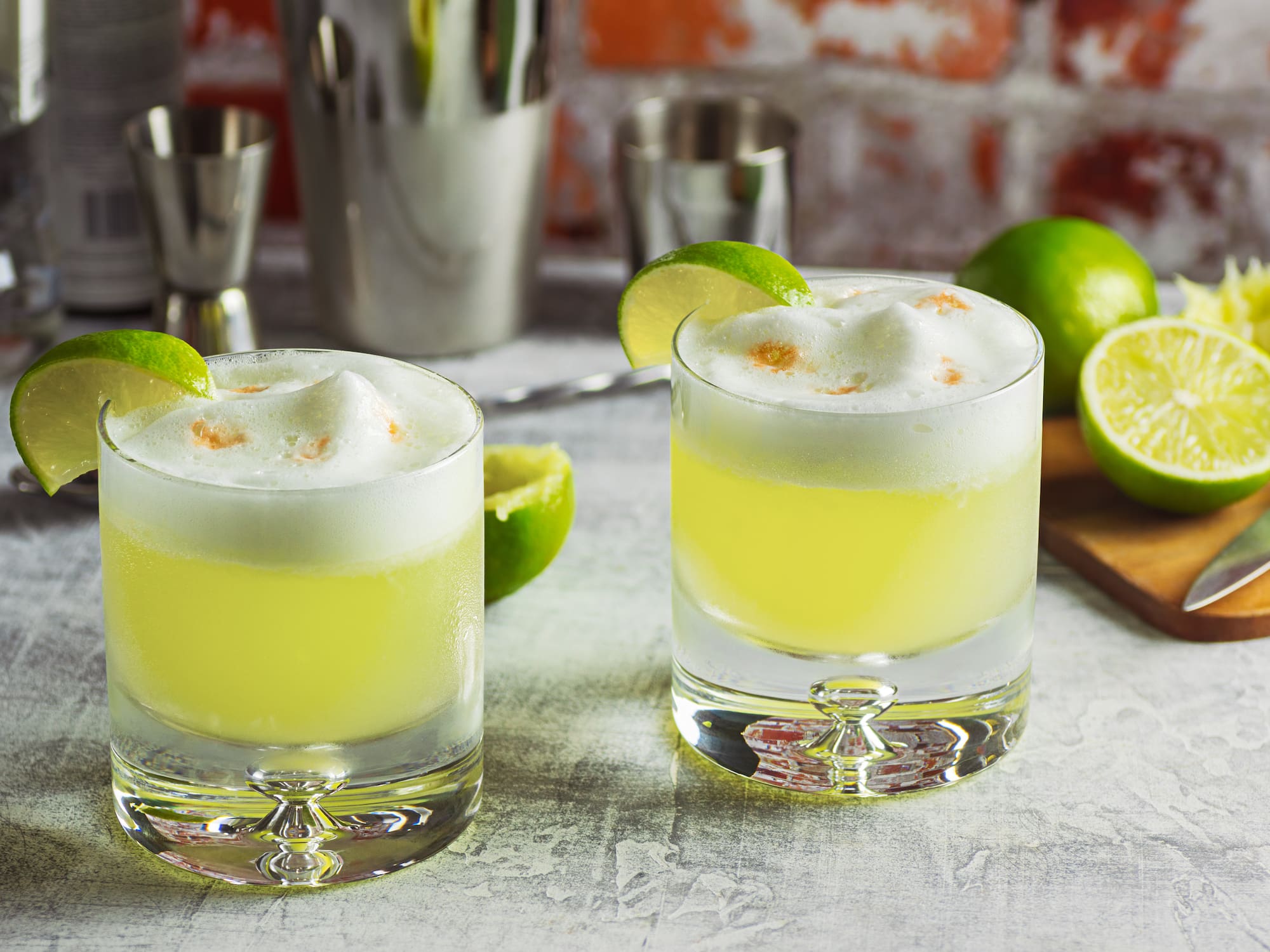 two glasses of yellow liquid with limes and a cutting board
