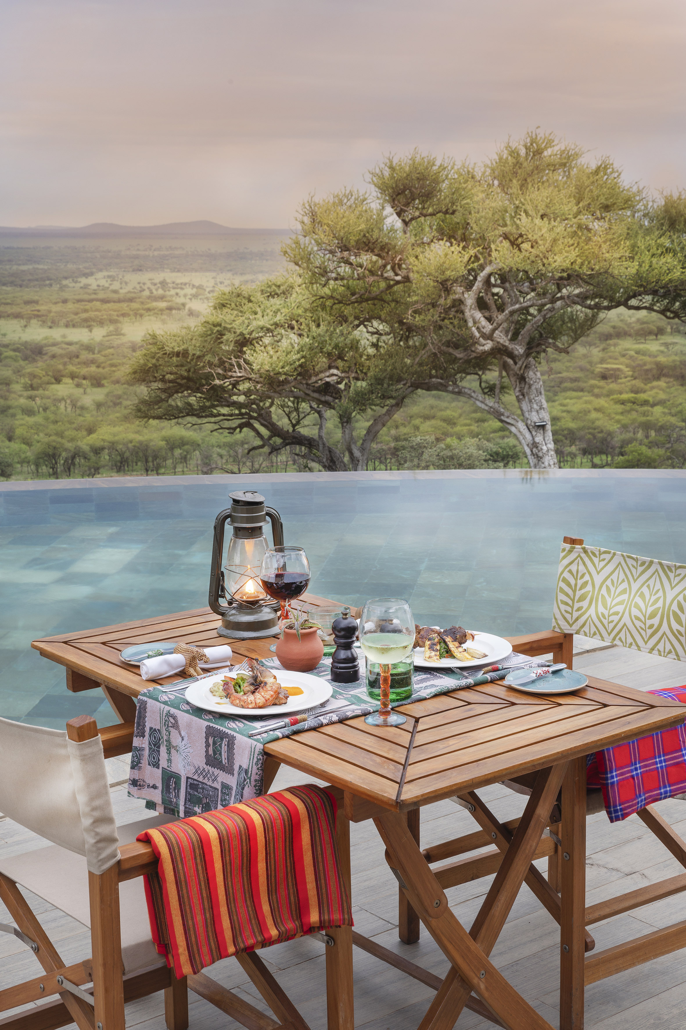Safari dinner for two by infinity pool, savanna landscape, lit lantern at dusk.