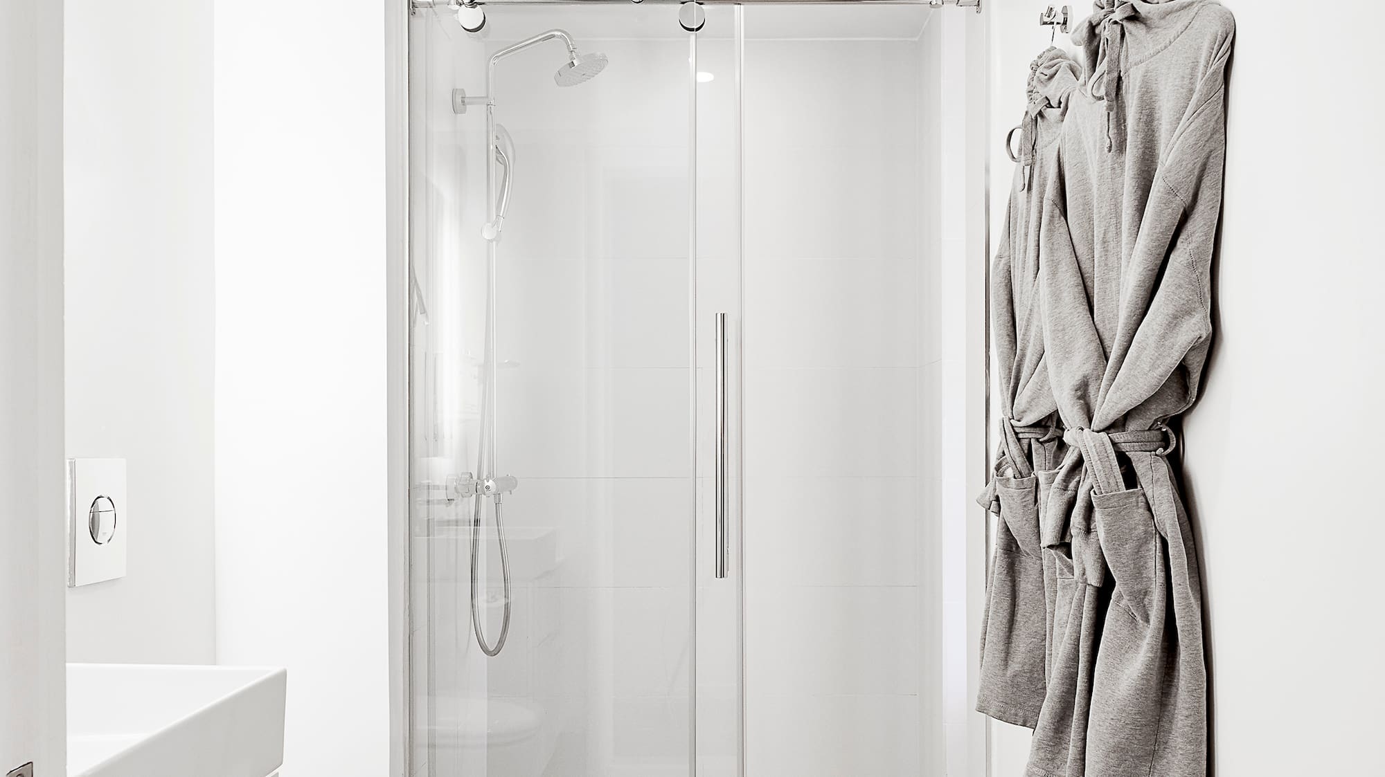 Modern white bathroom with glass shower, white vanity, gray tile floor, and bathrobes.