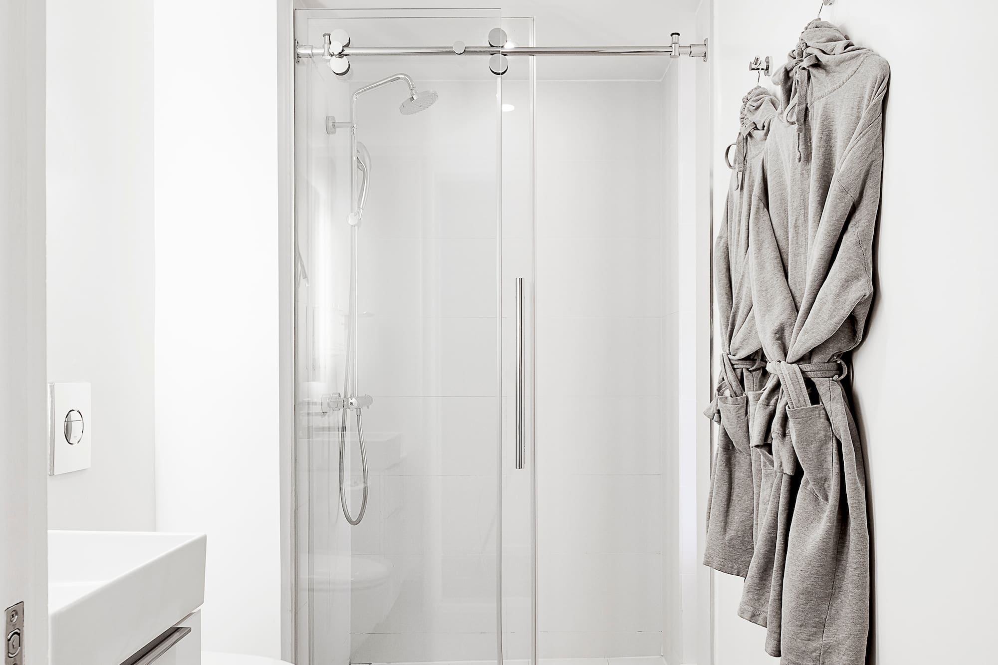 Modern white bathroom with glass shower, white vanity, gray tile floor, and bathrobes.