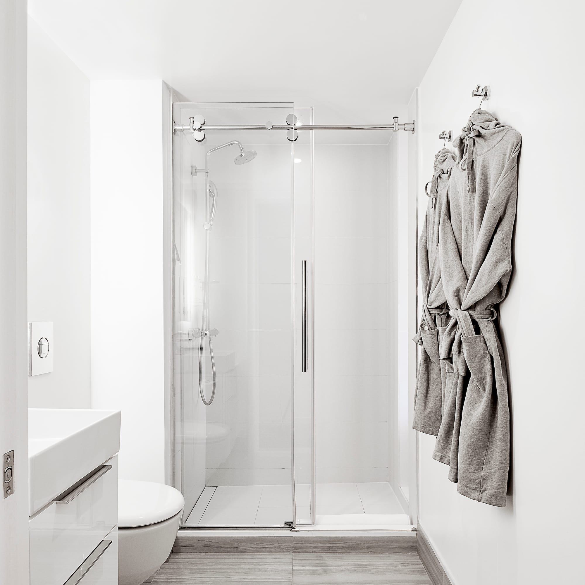 Modern white bathroom with glass shower, white vanity, gray tile floor, and bathrobes.
