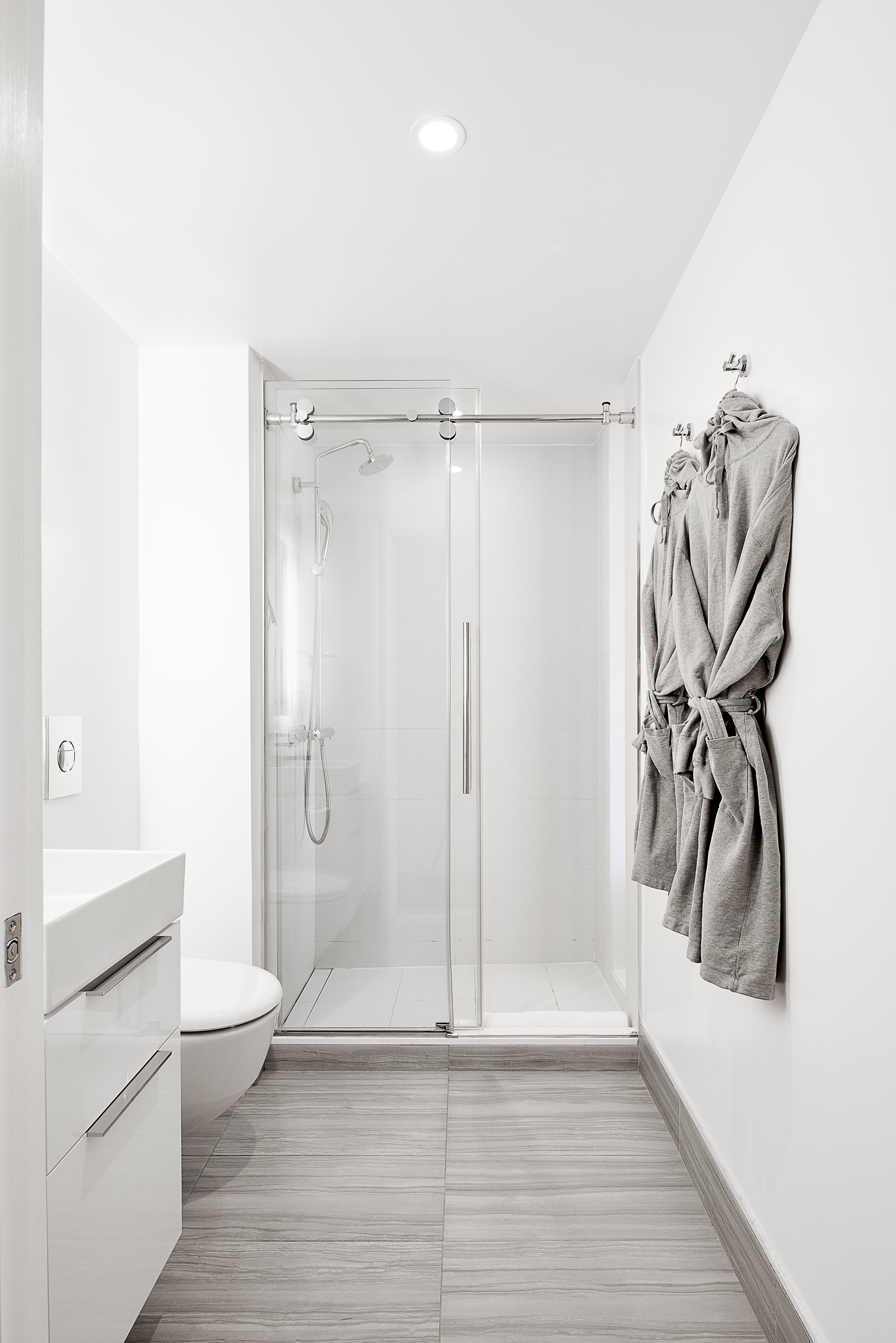 Modern white bathroom with glass shower, white vanity, gray tile floor, and bathrobes.
