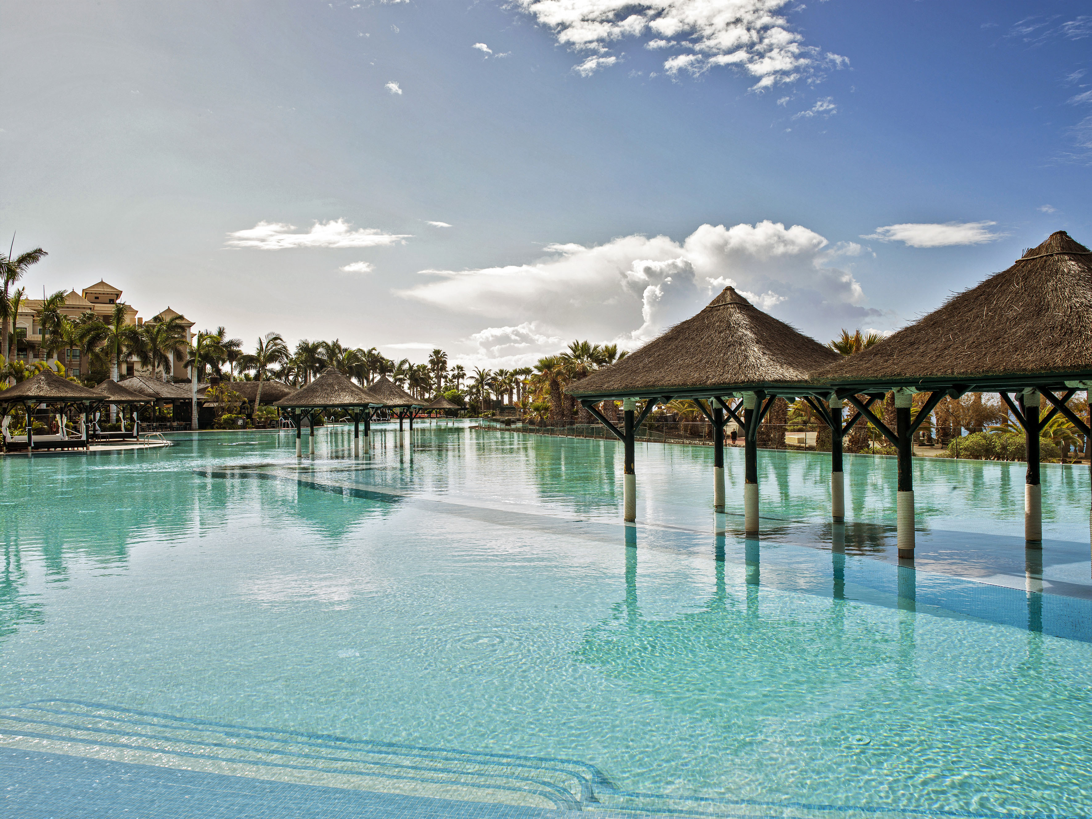 Tropical resort swimming pool with thatched cabanas and palm trees.
