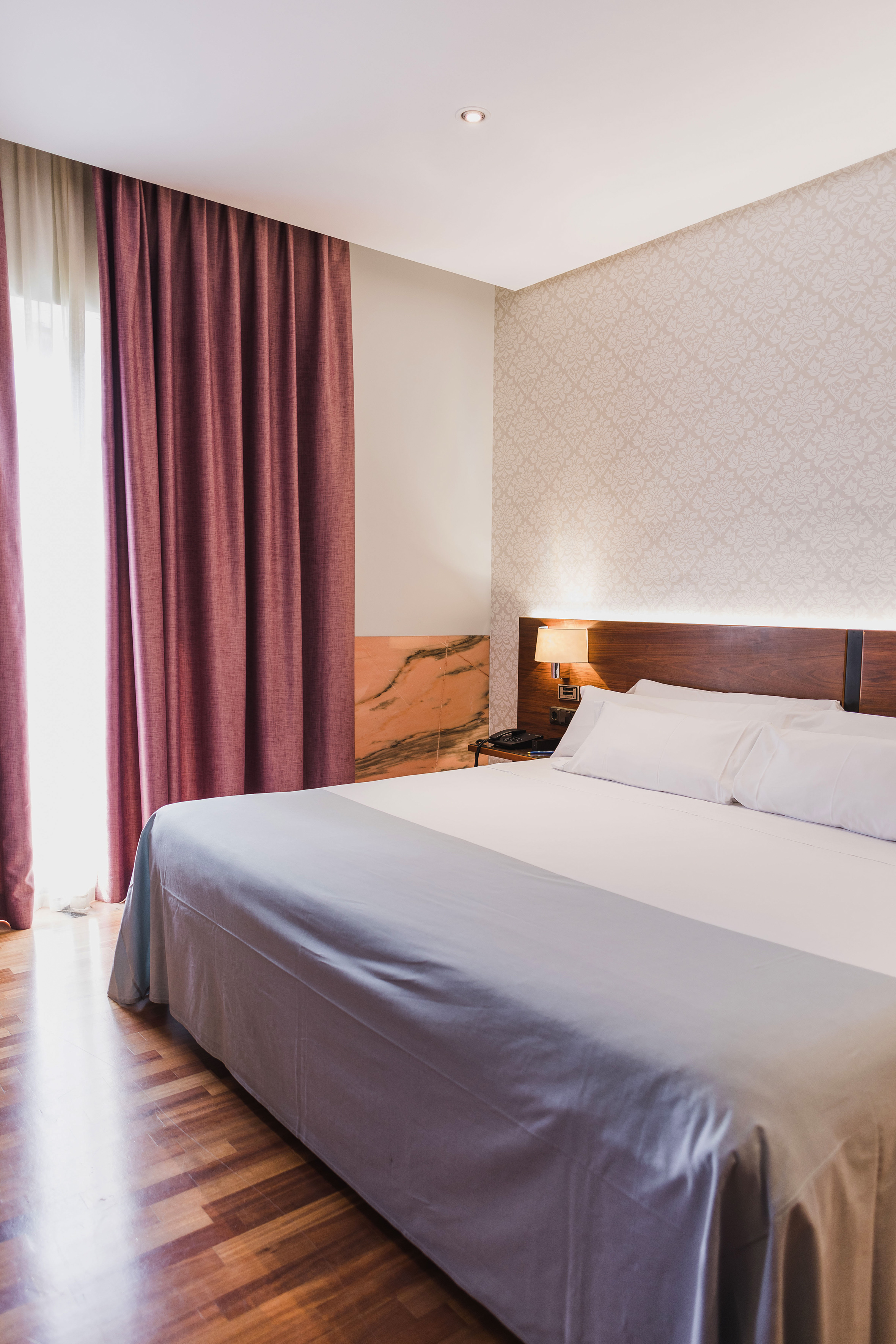 Modern hotel bedroom with bed, wooden headboard, patterned wall, plum curtains.