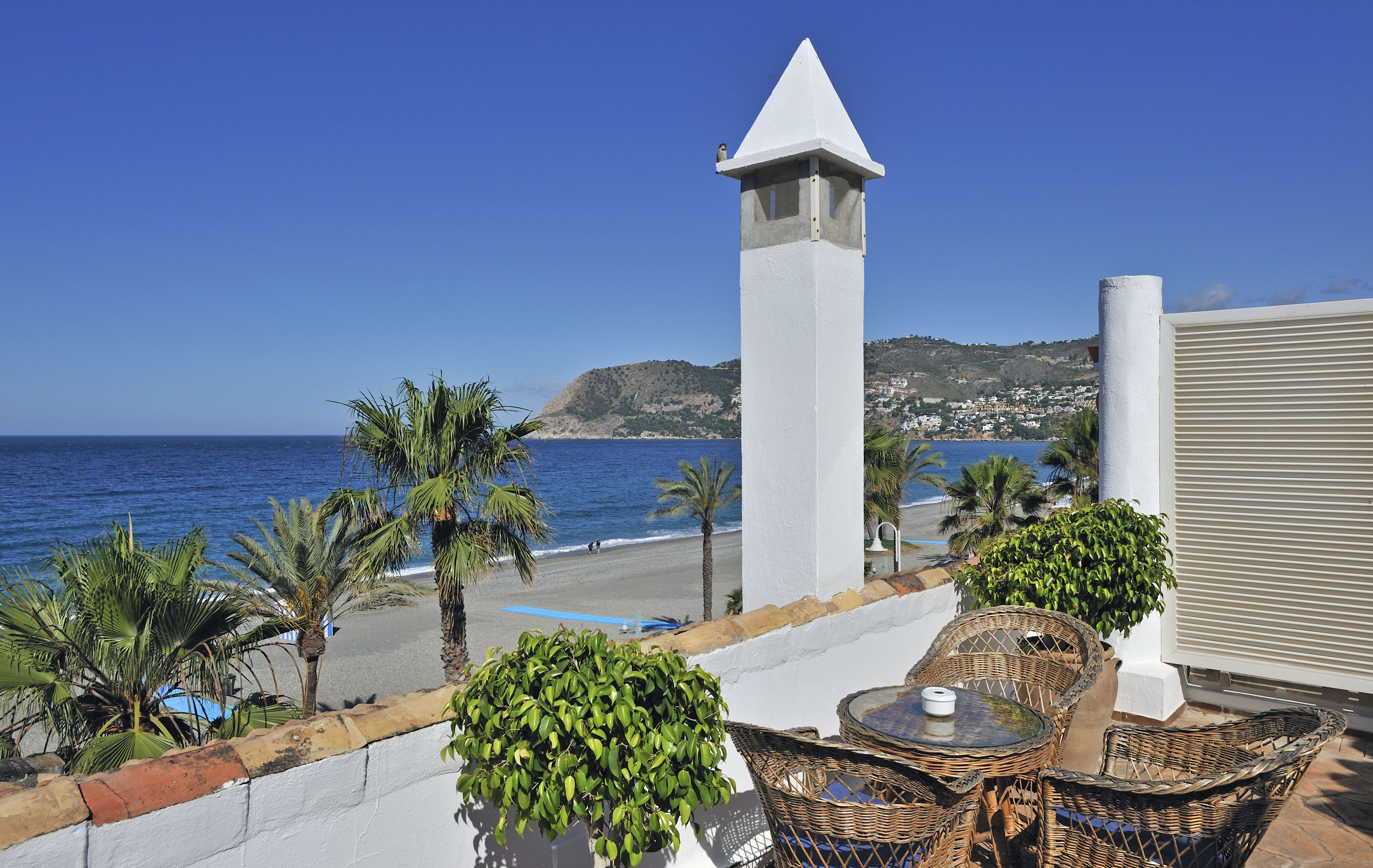 Coastal balcony view: sunny beach, blue sea, palm trees, mountains.