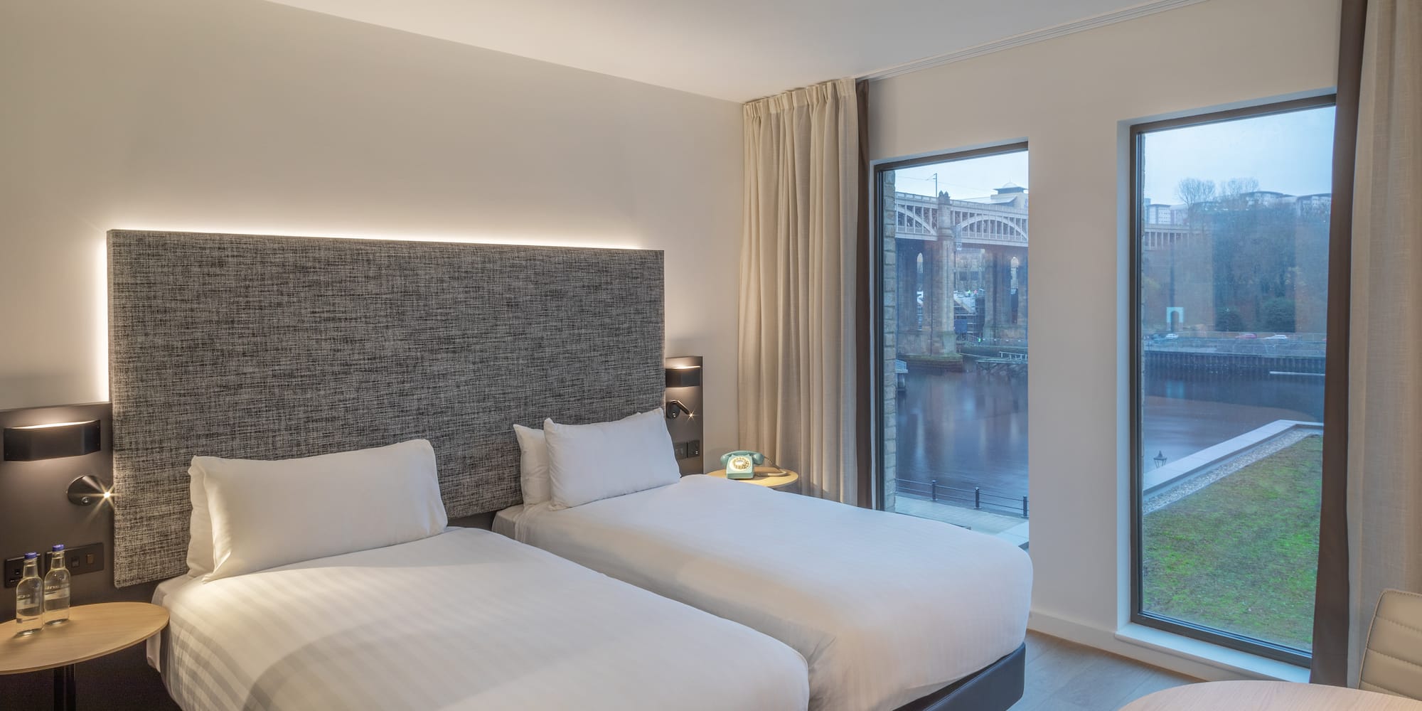 Modern hotel room with twin beds, river view, and bridge cityscape.