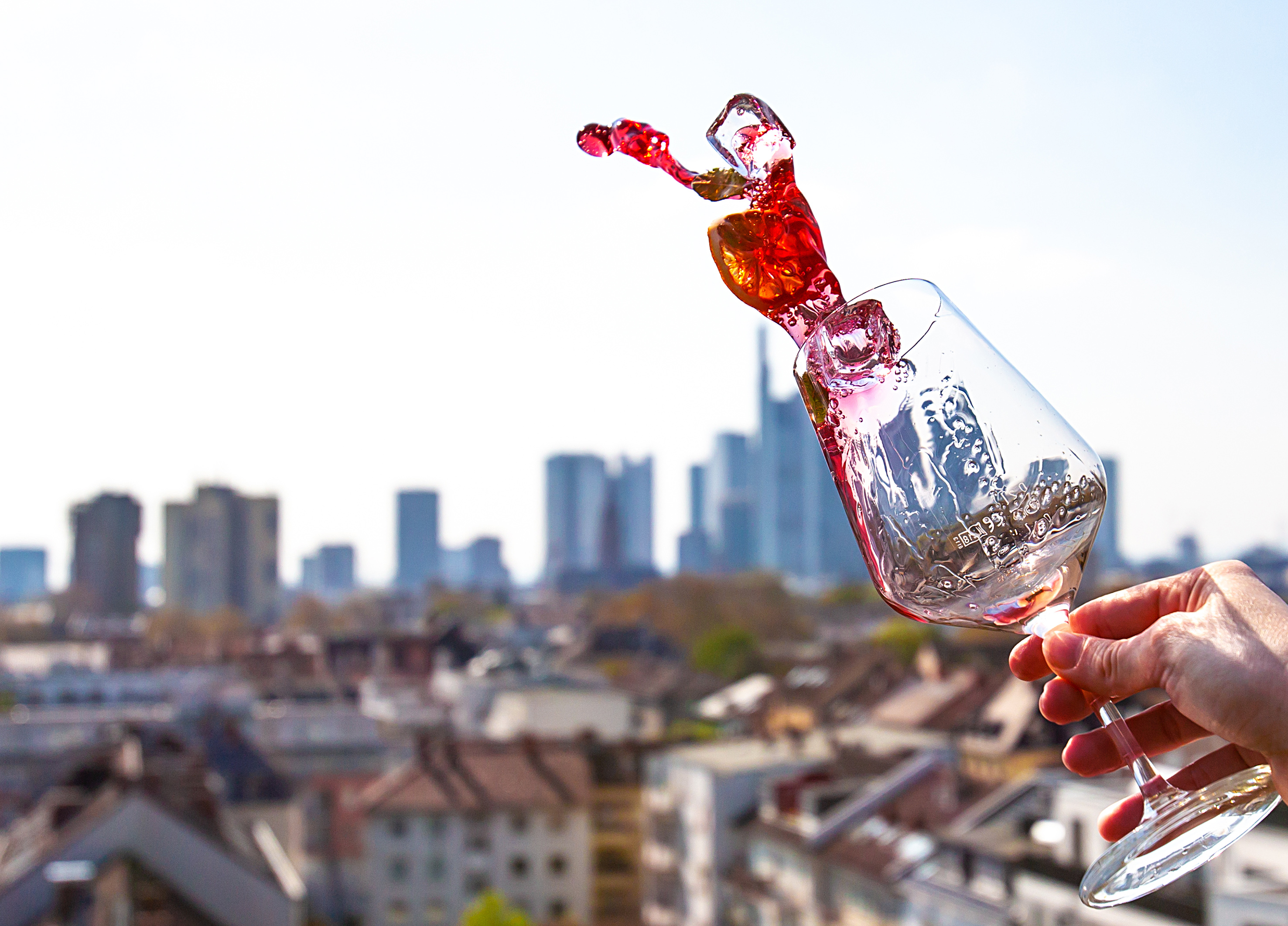 Red wine splashing from tilted glass, hand holding it, urban skyline.