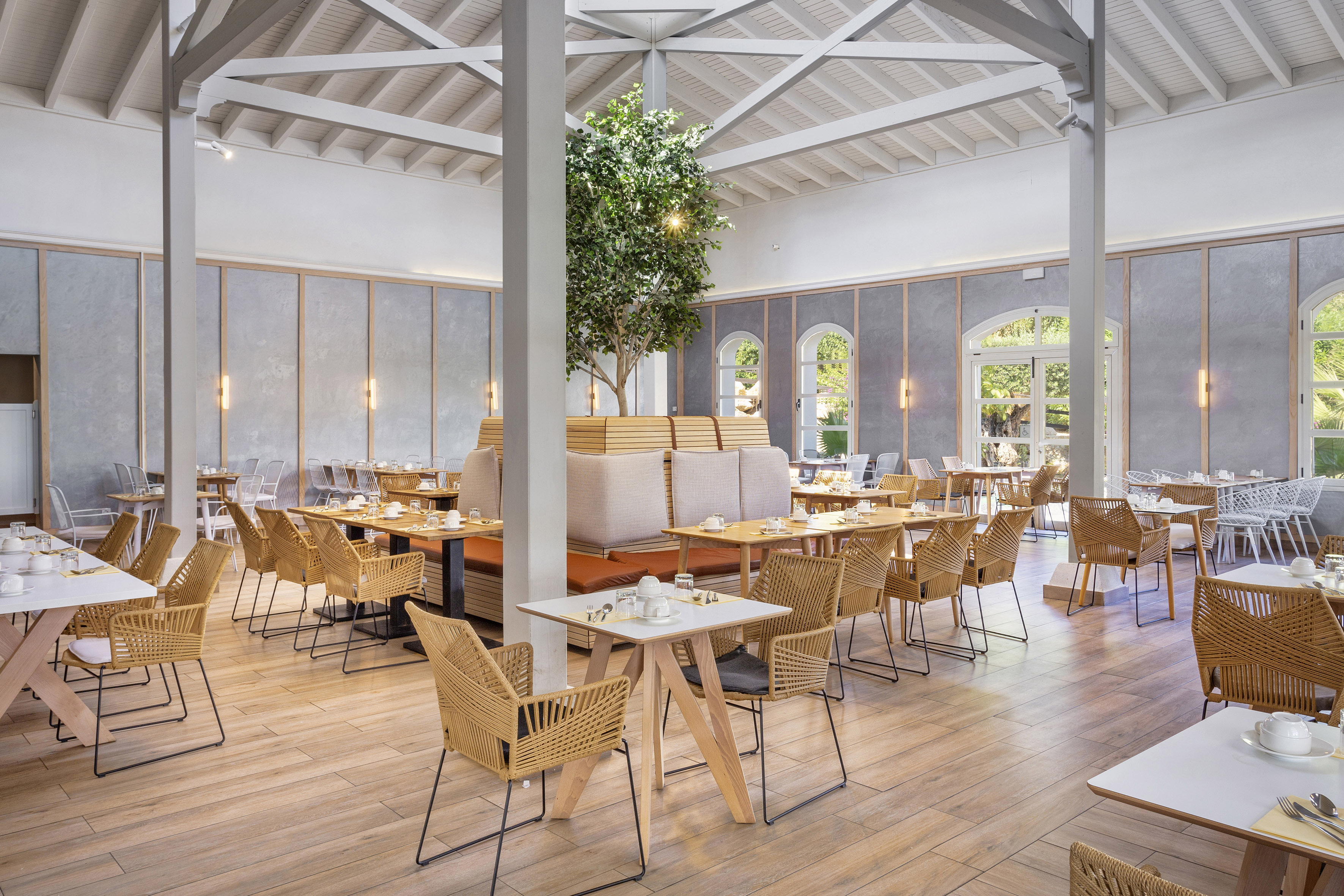 Modern hotel dining room, wicker chairs, light floor, vaulted ceiling, central tree.