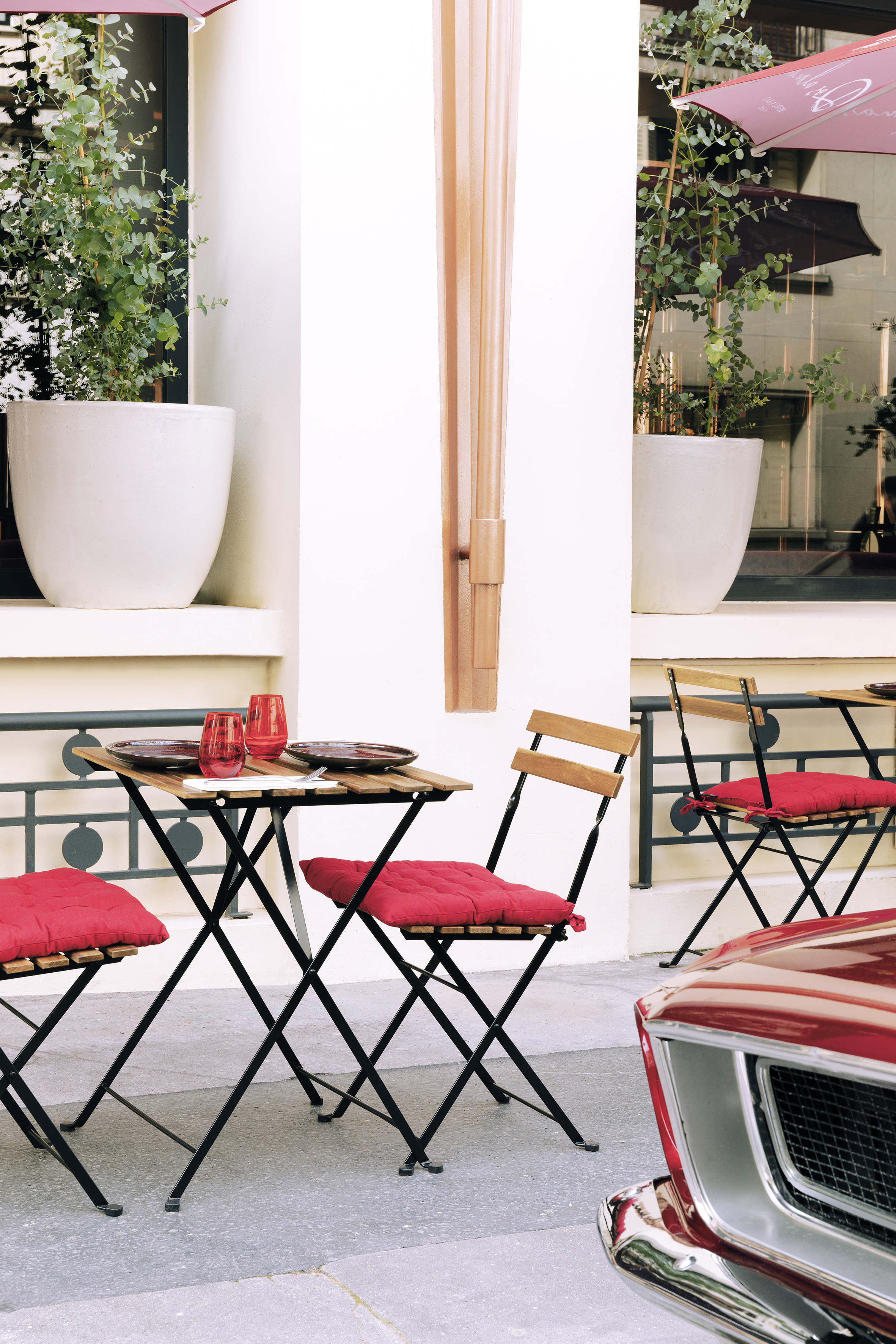 Outdoor bistro set with red cushions, tableware, potted plants, and red car.