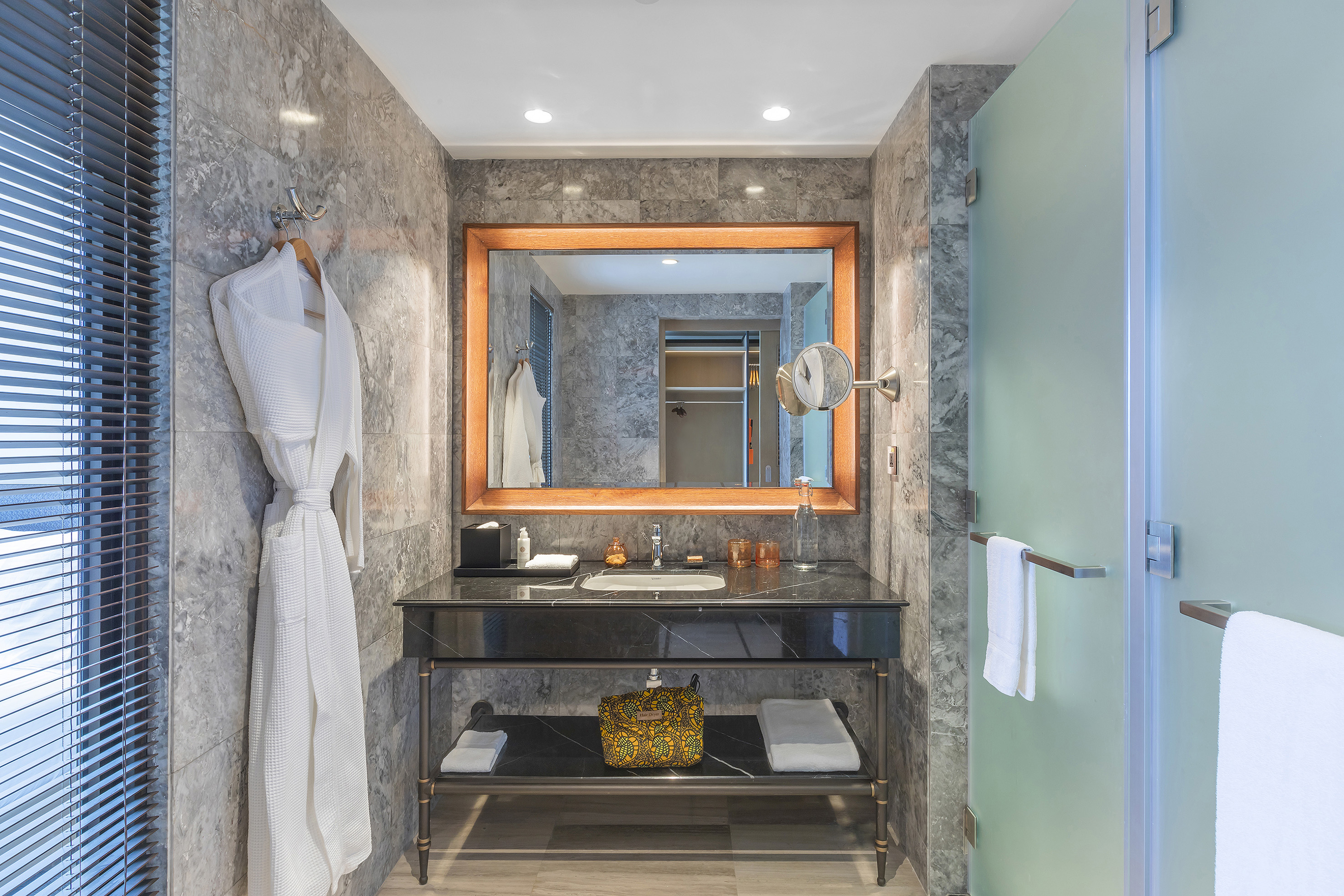 Modern hotel bathroom with marble vanity, mirror, white bathrobe, and frosted glass shower.