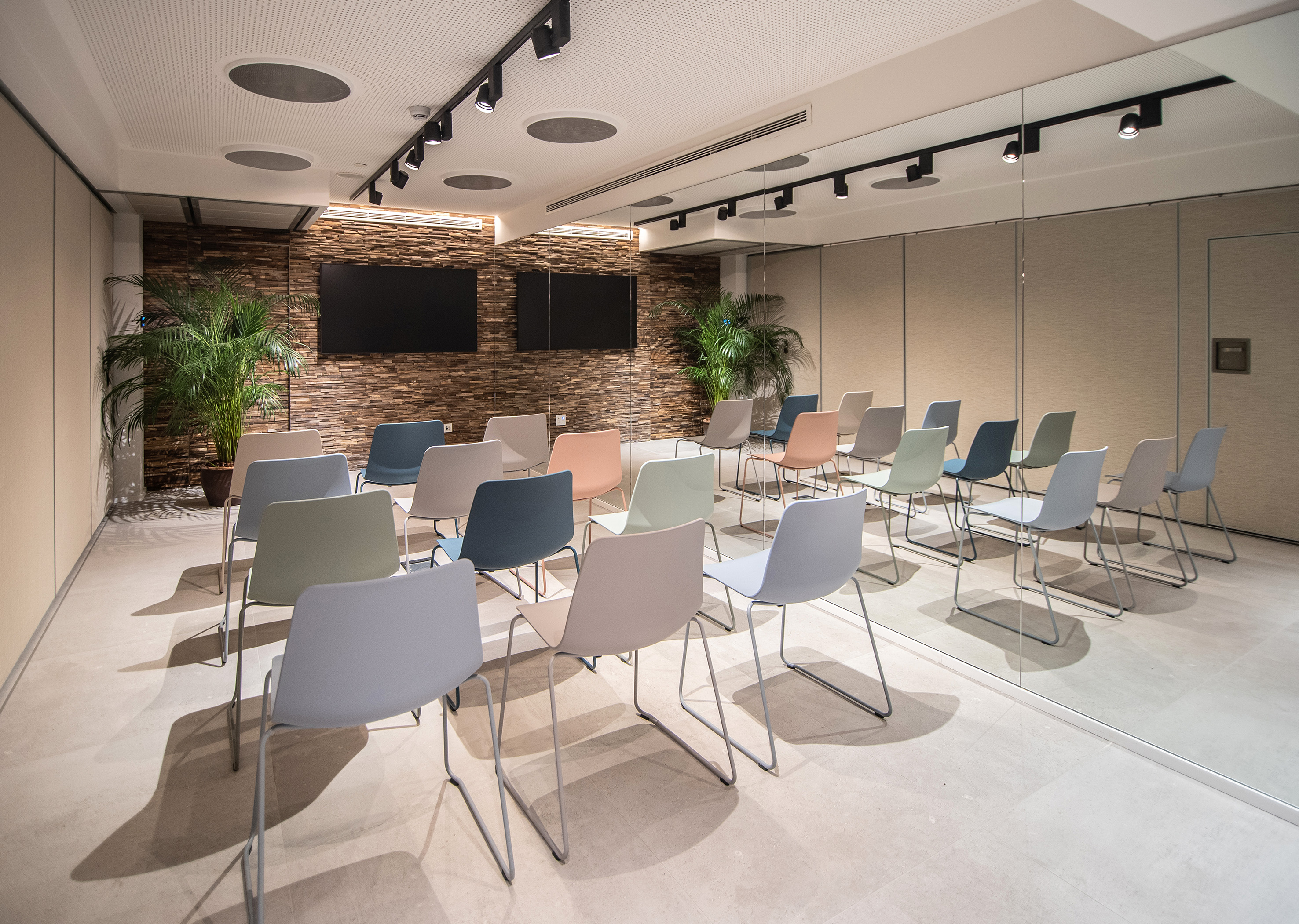 Modern conference room with colorful chairs, two screens, and a textured brick wall.
