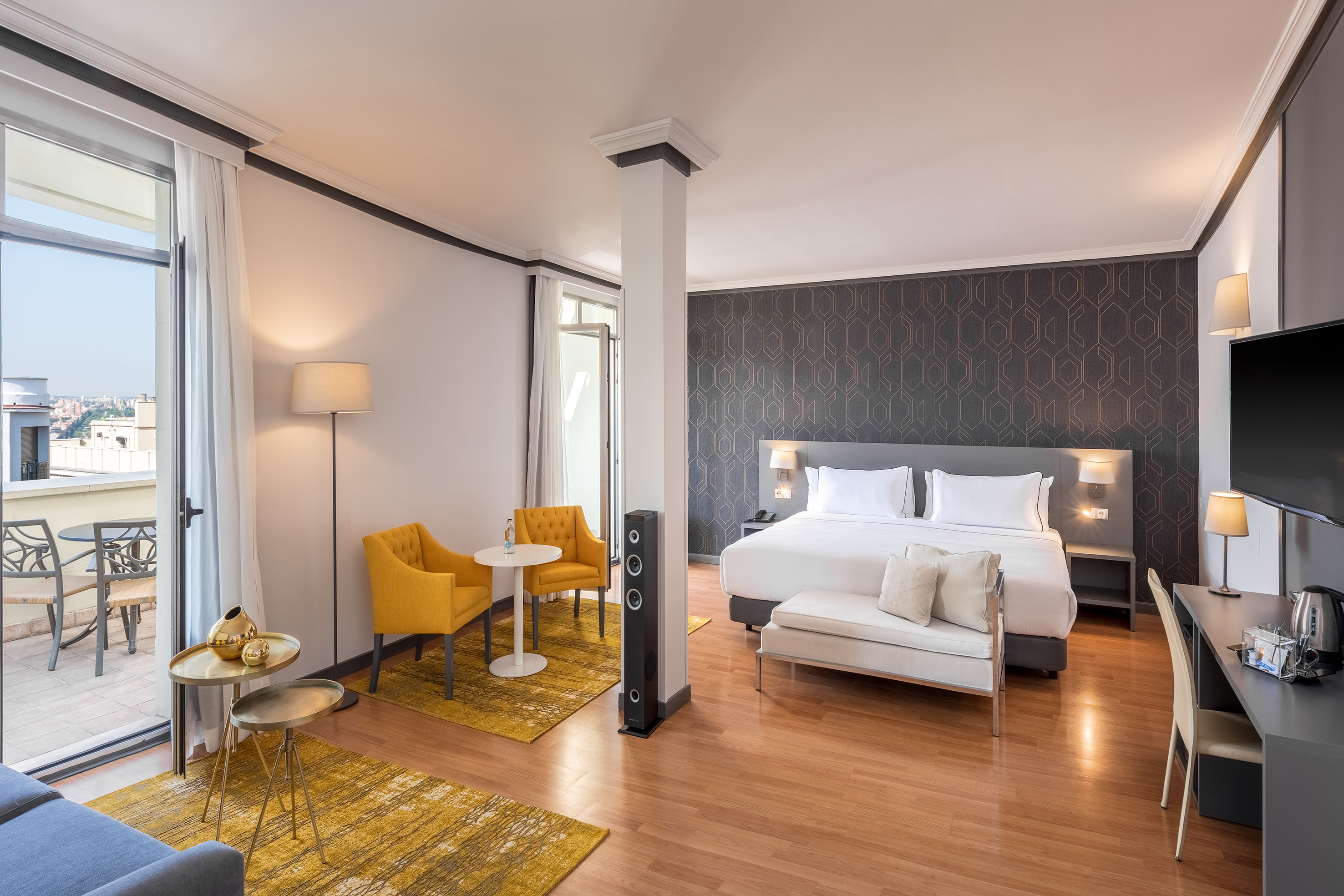 Hotel suite with large bed, balcony, yellow chairs, and modern furnishings.