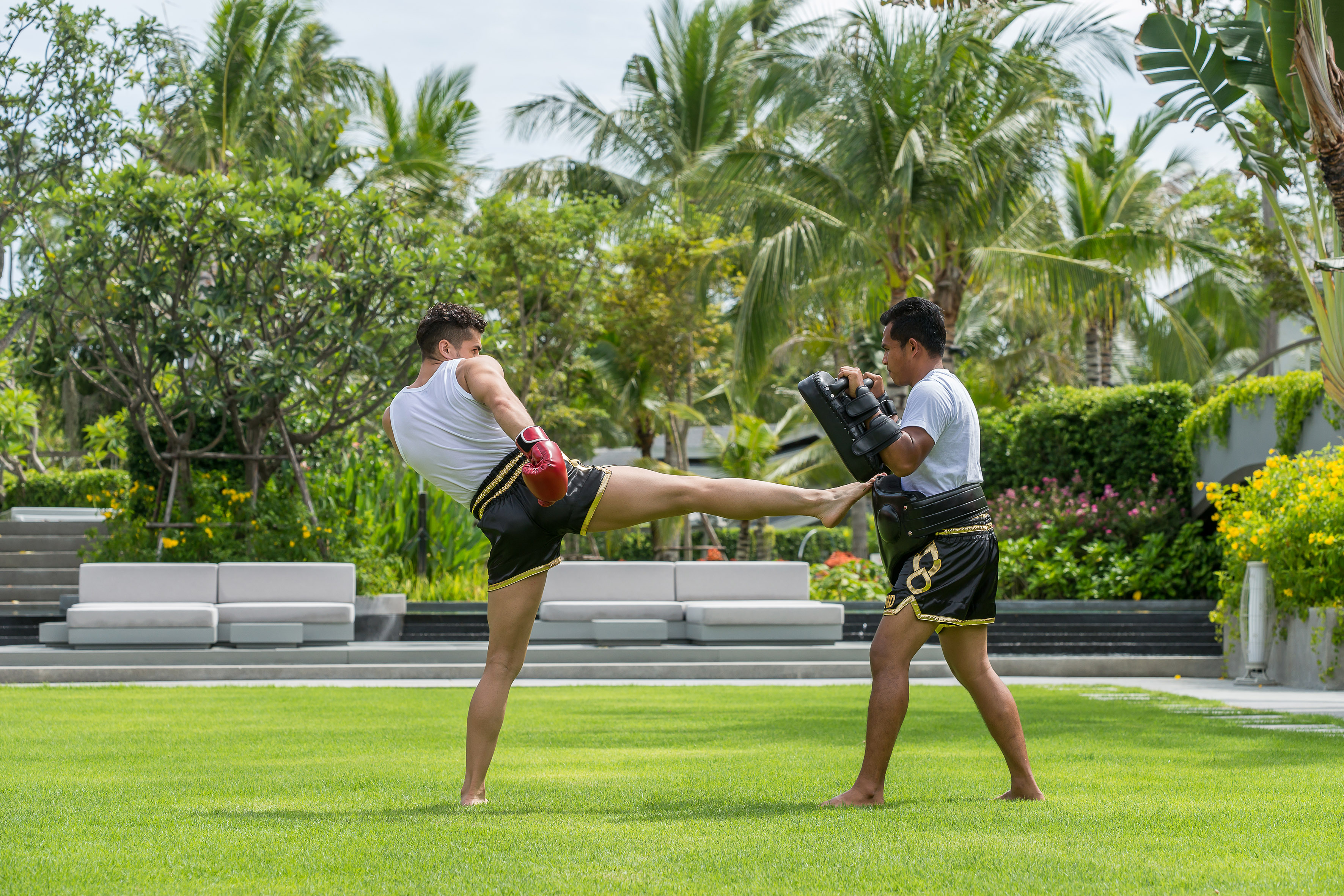 Two men doing Muay Thai kickboxing training on a tropical lawn.