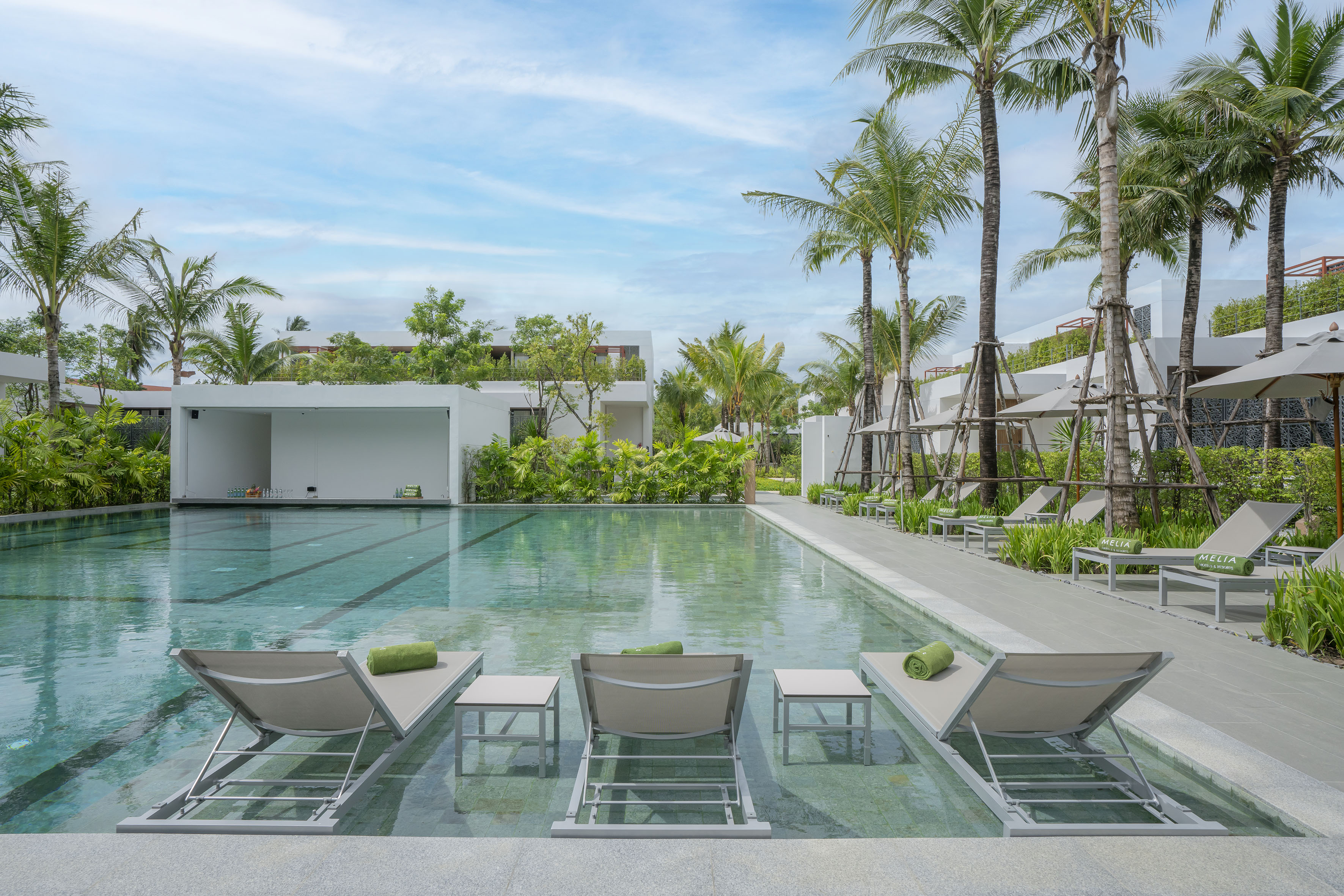 Tropical resort pool with in-water loungers, palm trees, and modern white buildings.